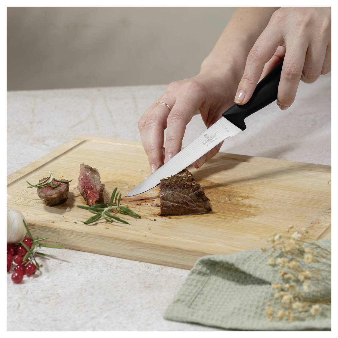 Hände schneiden gekochtes Steak auf einem hölzernen Schneidebrett mit einem Kochmesser, mit Kräutern und angerichtetem Dekor in der Nähe.