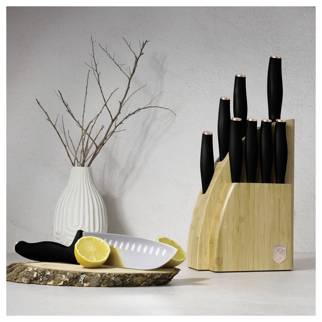 Holzblock für Messer mit schwarzen Messern neben einer weißen Vase mit Ästen; ein Messer mit Zitronen liegt auf einem hölzernen Tablett davor.