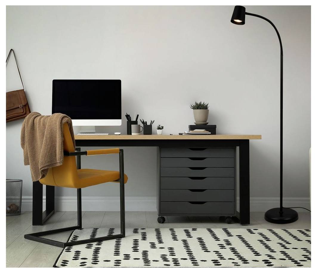 Minimales Homeoffice mit Schreibtisch, Computer, senfgelbem Stuhl, Stehleuchte und gemustertem Teppich vor einer schlichten Wand.