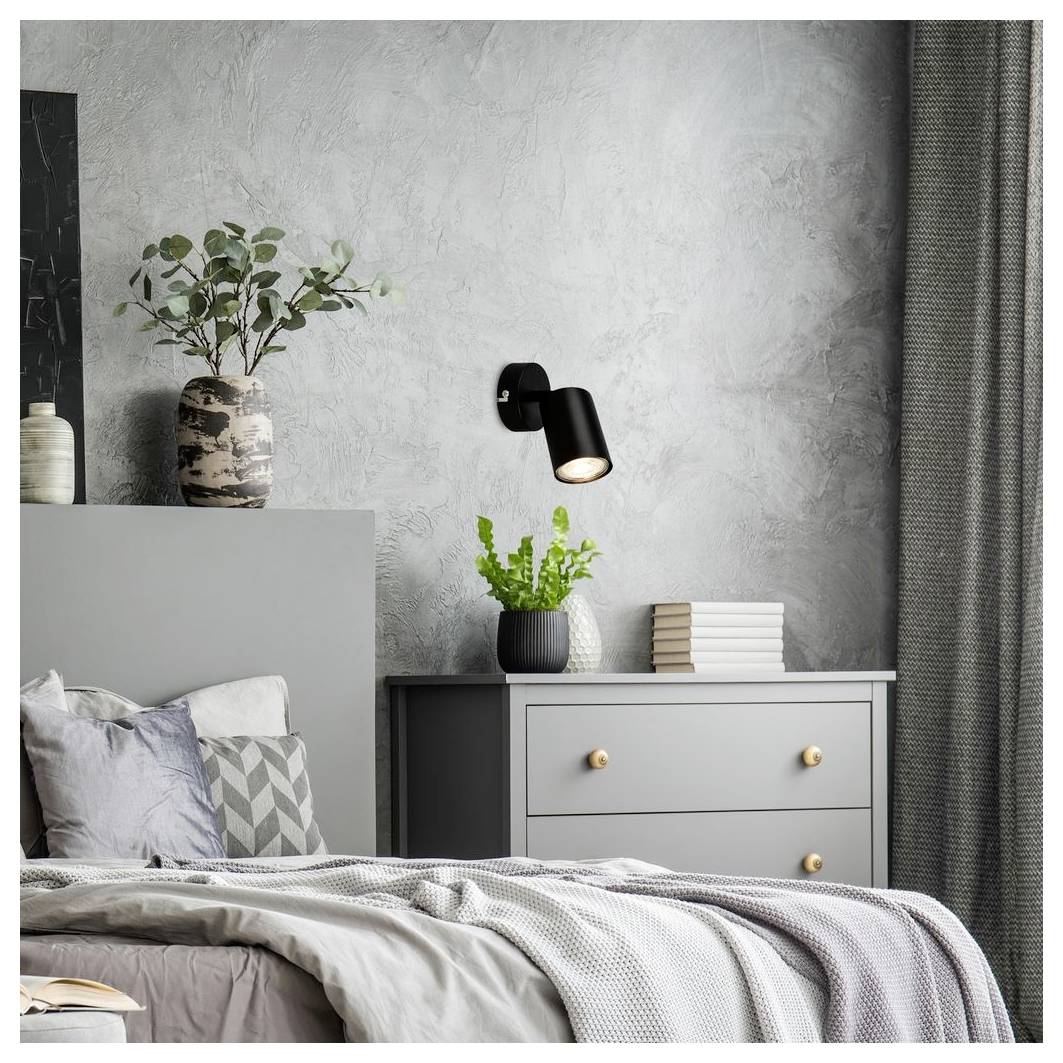 Modernes graues Schlafzimmer mit gemachtem Bett, Nachttisch, wandmontierter Lampe, Pflanzen und gestapelten Büchern gegen eine strukturierte Wand.