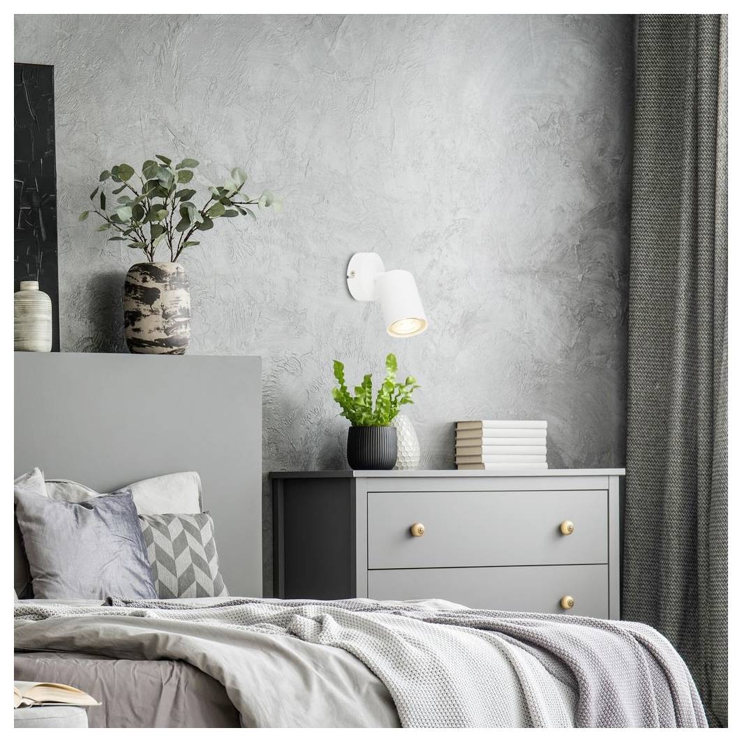 Modernes Schlafzimmer mit grauem Bett, Kissen, Kommode, Wandleuchte, Pflanzen, Büchern und Vorhängen vor einer strukturierten grauen Wand.
