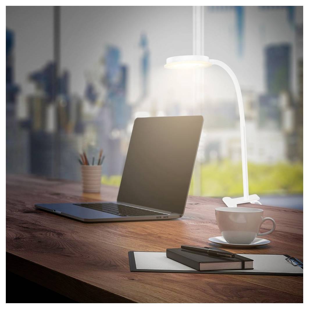 Laptop auf einem Holzschreibtisch neben einer Kaffeetasse, einer Lampe und Notizbüchern, mit einer unscharfen Stadtkulisse, die durch das Fenster sichtbar ist.