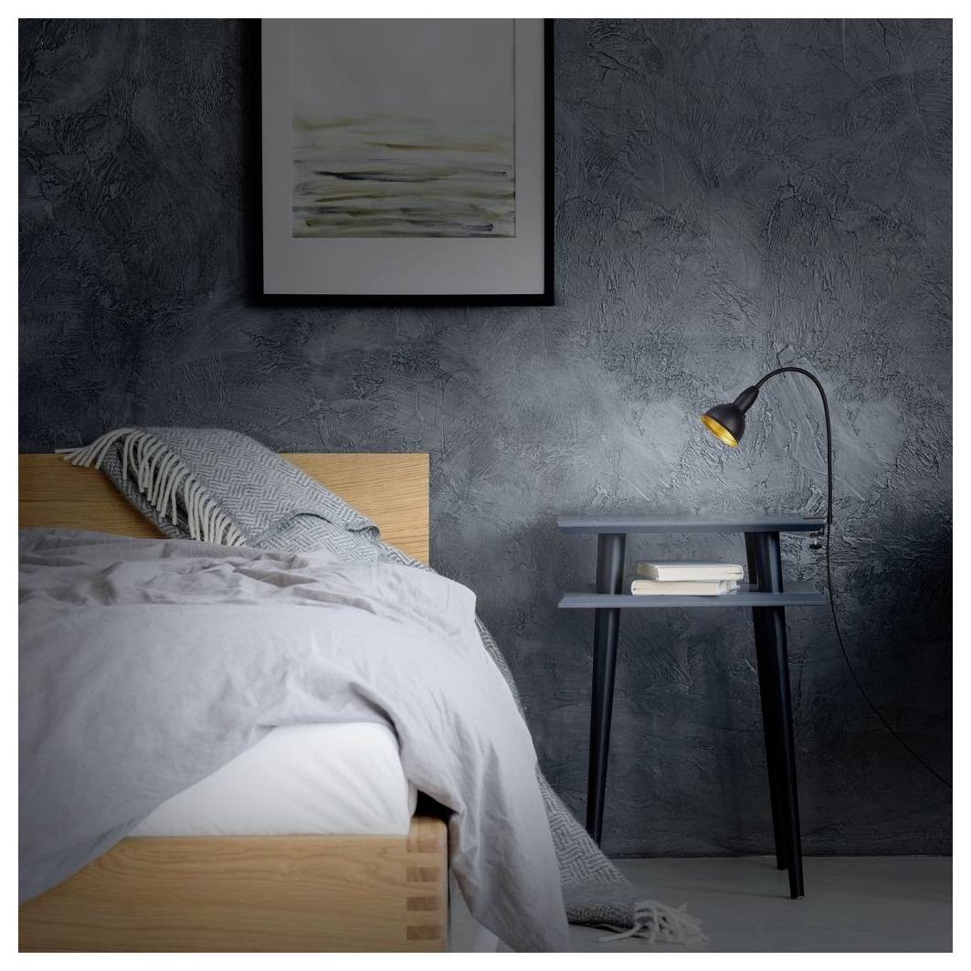 Ein ordentlich gemachtes Bett mit grauer Bettwäsche neben einem kleinen schwarzen Nachttisch und einer leuchtenden Lampe in einem dunklen, minimalistischen Schlafzimmer.