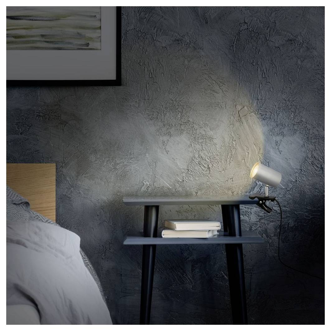 Nachttisch im Schlafzimmer mit einer leuchtenden Spotlightlampe, gestapelten Büchern und einem Teil eines Bettes vor einer dunkel strukturierten Wand.