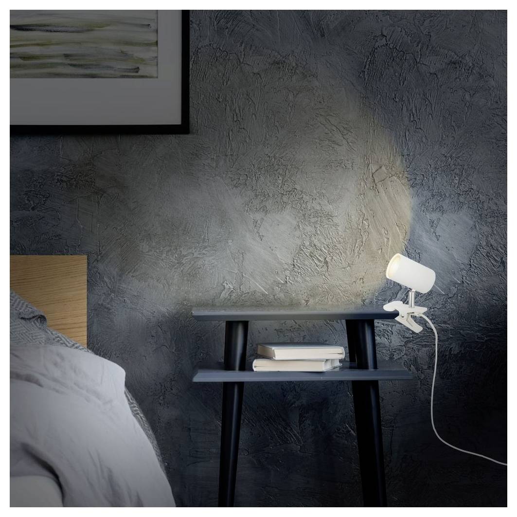 Düstere Schlafzimmerszene mit einem Nachttisch, gestapelten Büchern und einer leuchtenden verstellbaren Lampe vor einer strukturierten grauen Wand neben einem Bett.
