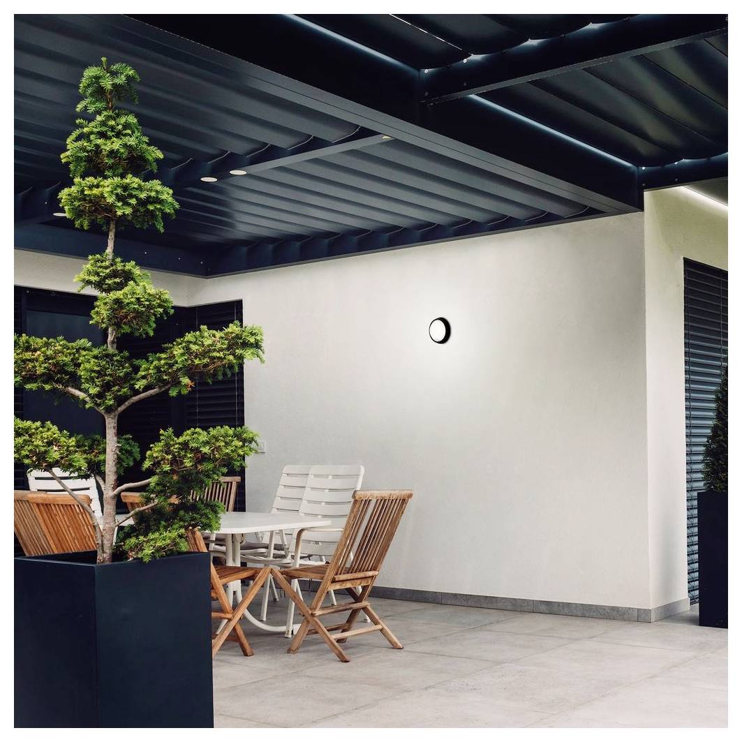Modernes Patio mit Topfbaum, weißem Tisch und Stühlen sowie einer dunklen Lamelldecke neben einer schlichten Wand mit einer runden Leuchte.