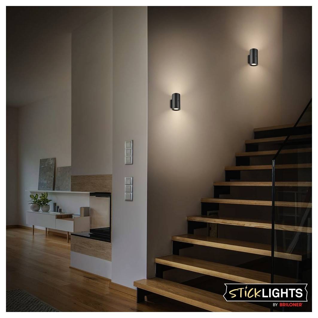 Moderne Treppe mit zwei Wandleuchten, die Licht auf eine braune Wand werfen, neben einem offenen Wohnbereich mit Holzböden und einem Kamin.