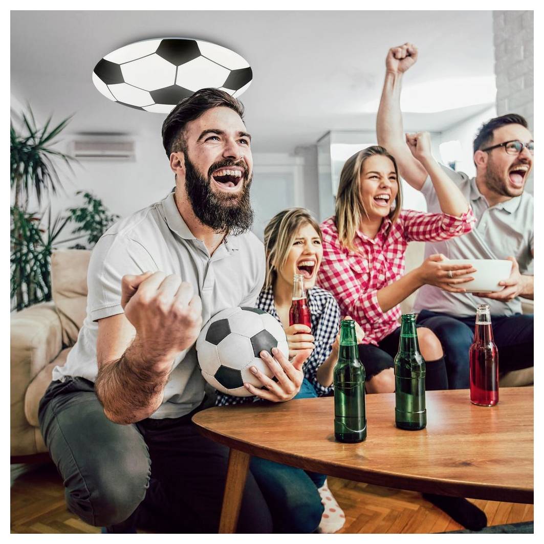 Freunde jubeln zu Hause, während sie ein Fußballspiel schauen, ein Mann hält einen Ball und die anderen feiern auf dem Sofa mit Getränken in der Nähe.