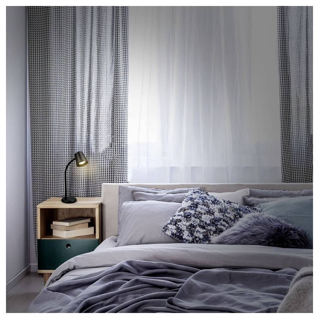 Ordentlich gemachtes Bett mit grauer Bettwäsche und dekorativen Kissen neben einem kleinen hölzernen Nachttisch und einer Lampe in einem sanft beleuchteten Schlafzimmer.