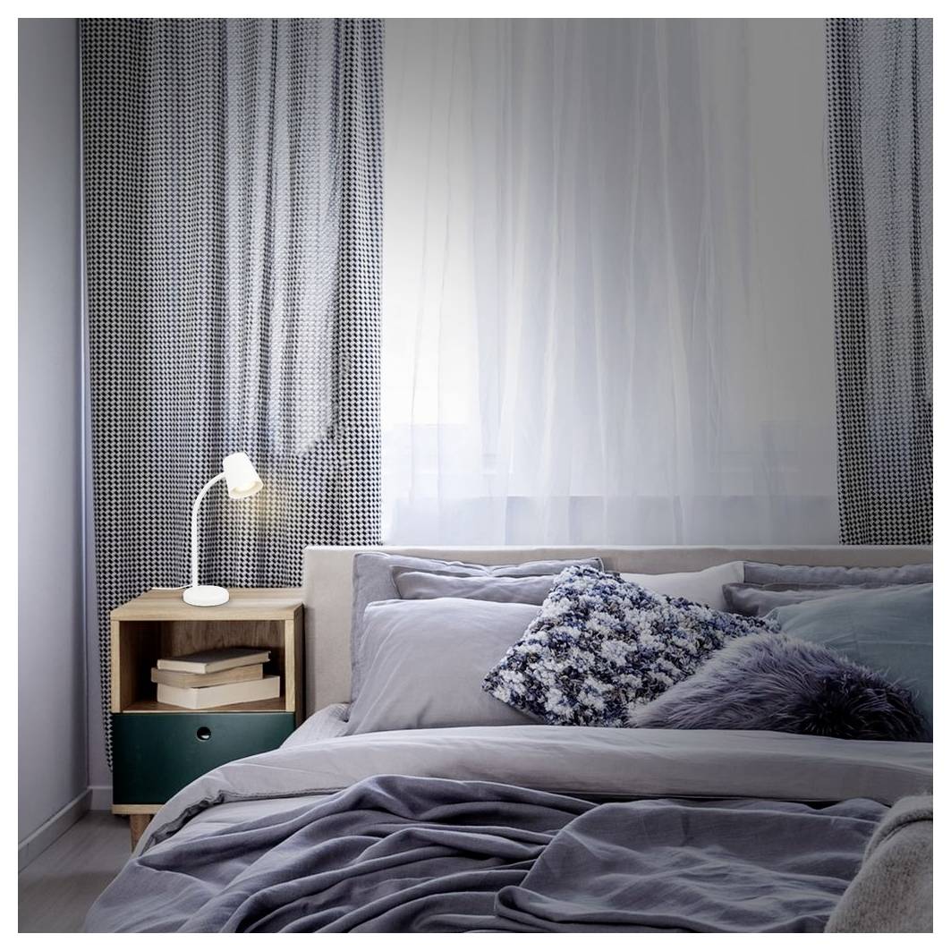 Ordentlich gemachtes Bett mit grauer Bettwäsche und dekorativen Kissen neben einem kleinen hölzernen Nachttisch und einer Lampe in einem sanft beleuchteten Schlafzimmer.