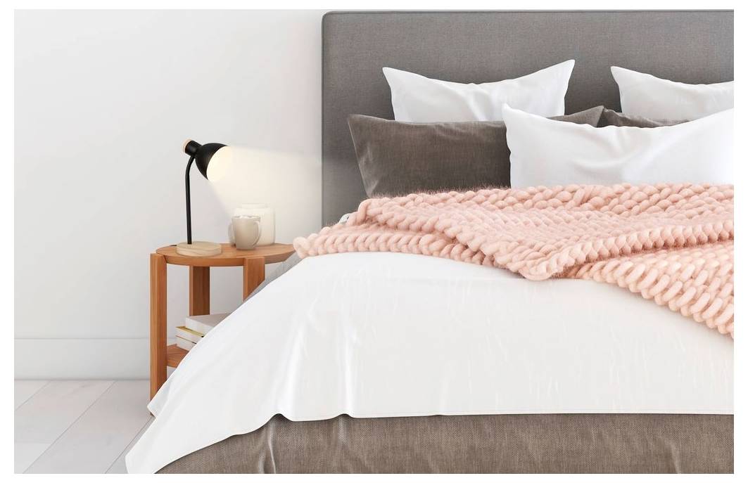 Ordentlich gemachtes Bett mit grauem Kopfteil, weißer Bettwäsche, rosa Wurfdecke und einem Nachttisch mit Lampe.