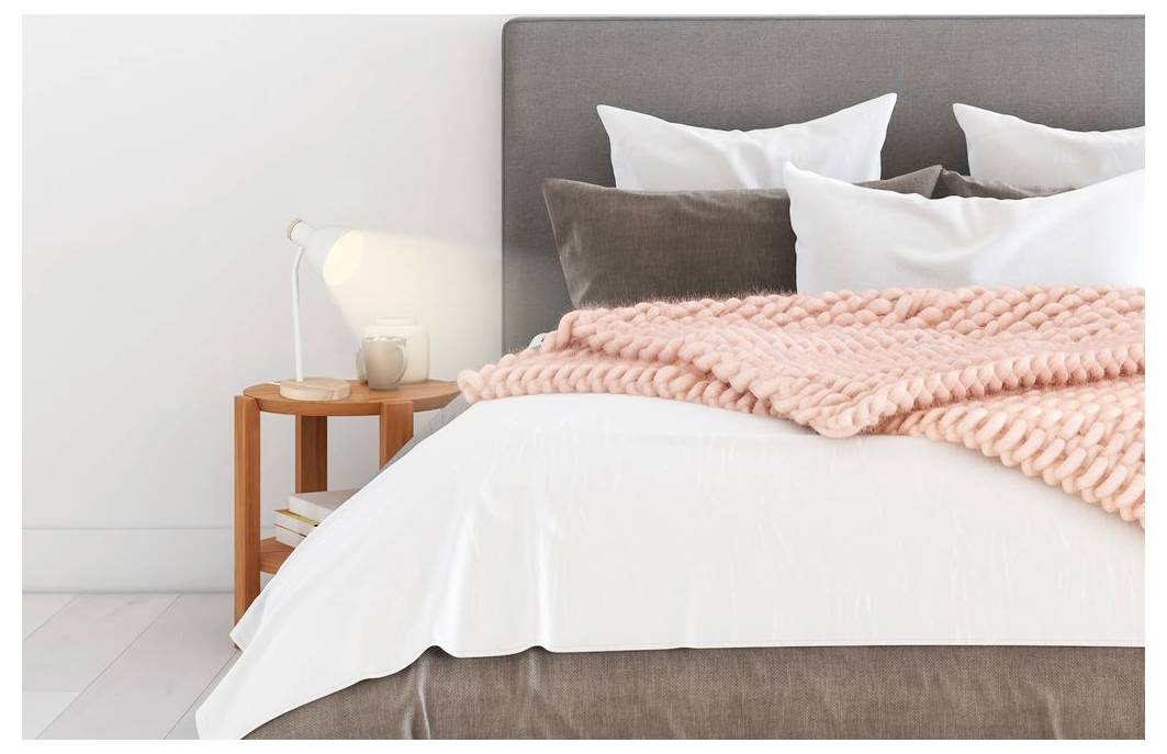 Ein ordentliches Bett mit weißer Bettwäsche, braunem Kopfteil und einer rosa gestrickten Wurfdecke in einem hellen Schlafzimmer neben einem Holznachttisch und einer Lampe.
