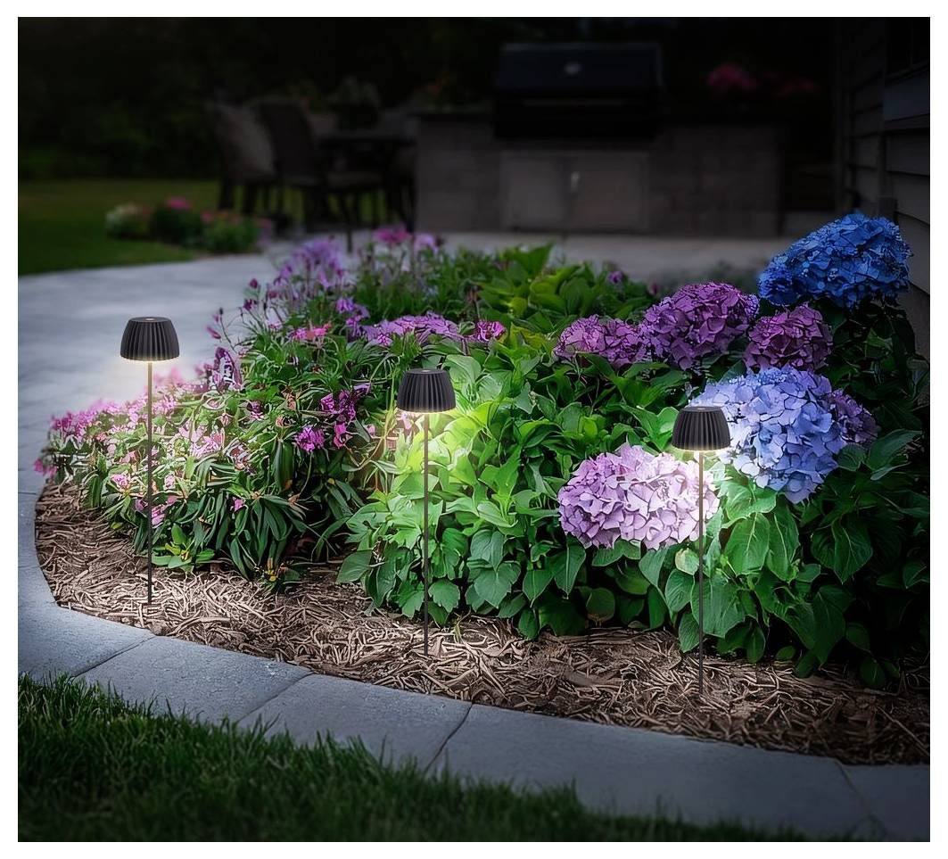 Drei kleine Gartenwegleuchten beleuchten ein Blumenbeet mit farbenfrohen Hortensien und rosa blühenden Pflanzen entlang einer geschwungenen Bordsteinkante in der Dämmerung.