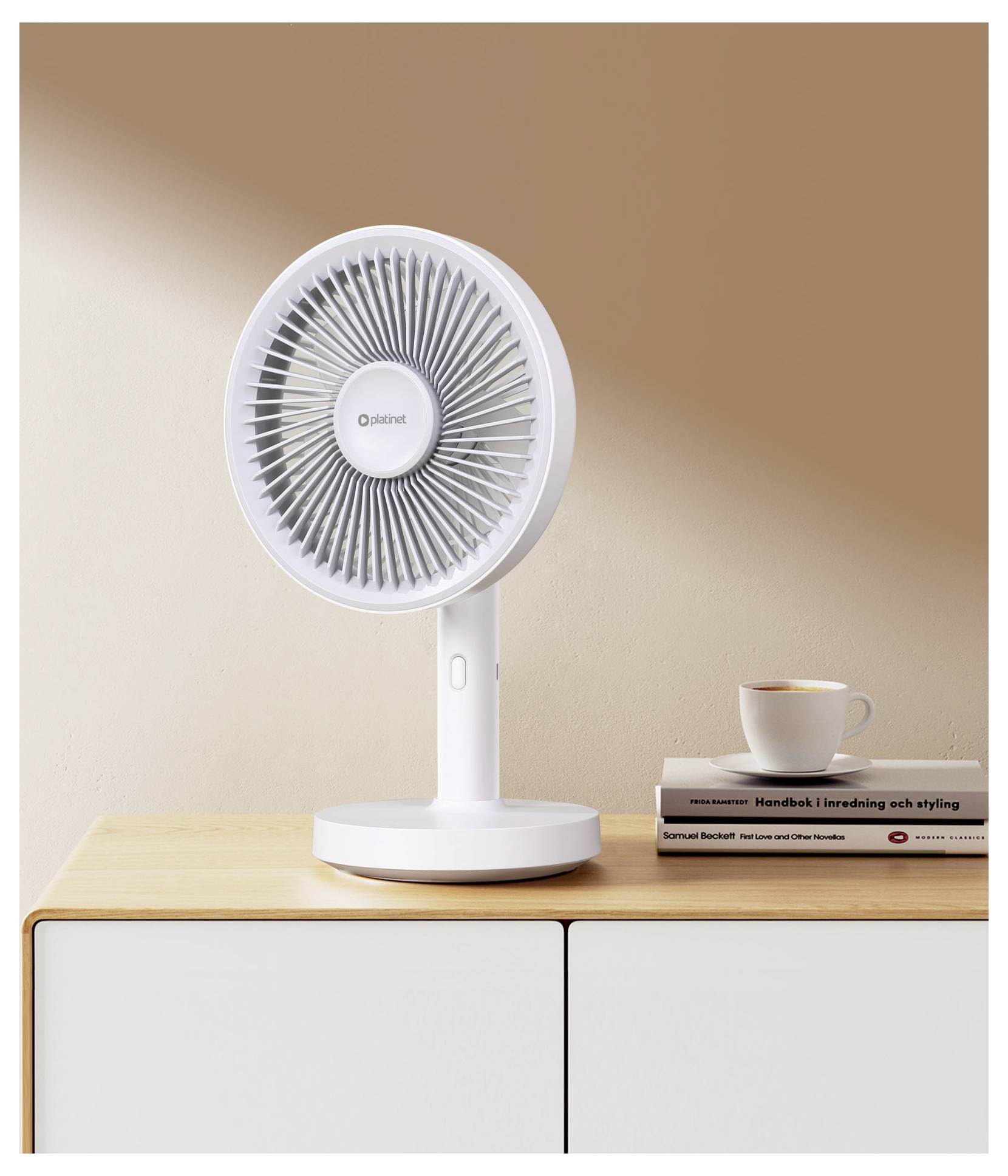 Weißer Tischventilator auf einem Holzschrank neben gestapelten Büchern und einer Kaffeetasse vor einer beigen Wand.