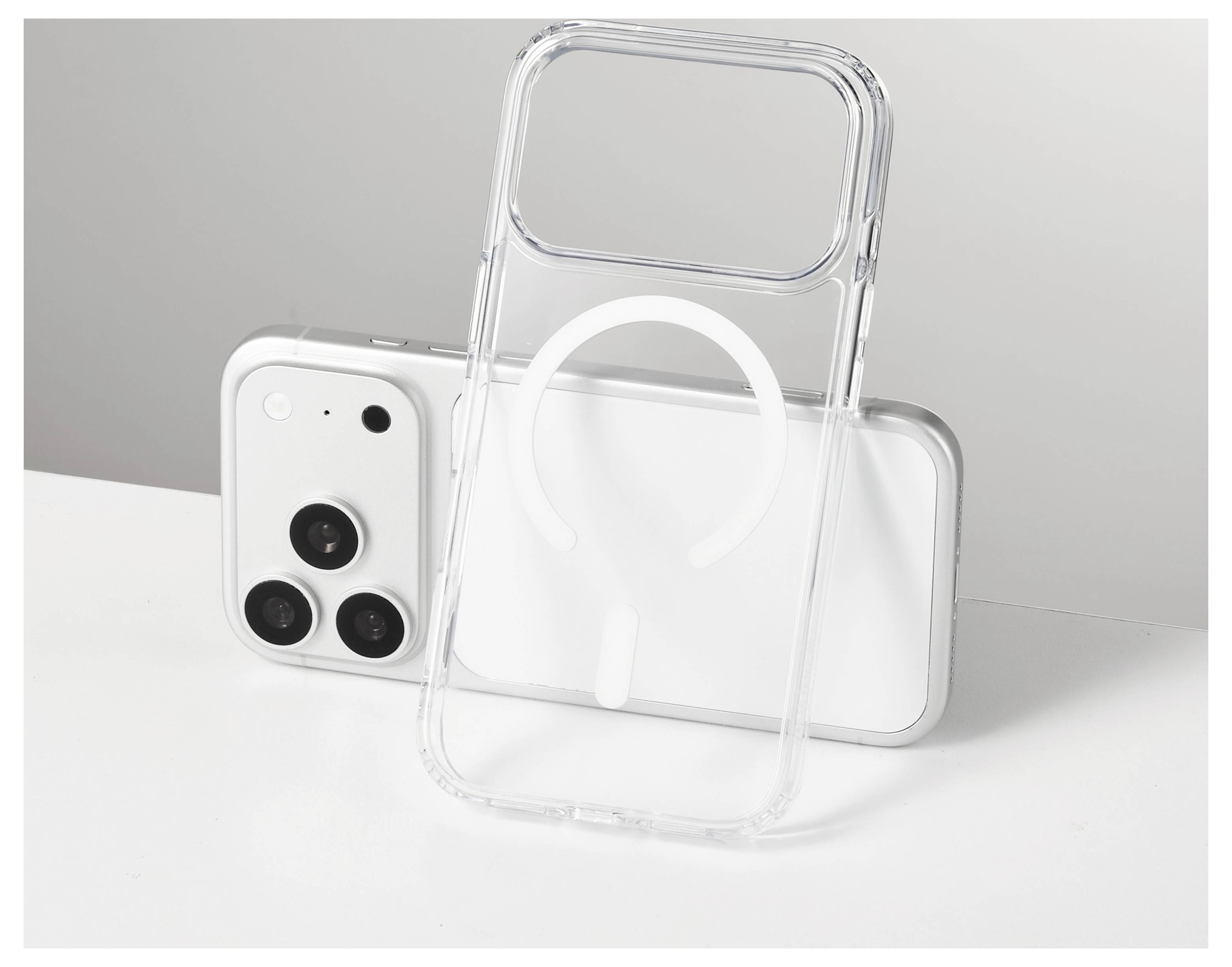 Durchsichtige Handyhülle lehnt an einem weißen Smartphone, zeigt einen großen MagSafe-ähnlichen Ring auf einer minimalistischen Tischplatte.