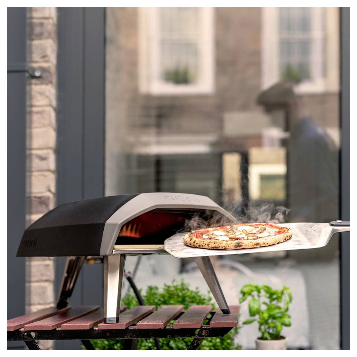 Pizza wird mit einer Schaufel aus einem Holzfeuer-Außenofen auf einem Terrassentisch herausgenommen, mit aufsteigendem Rauch.