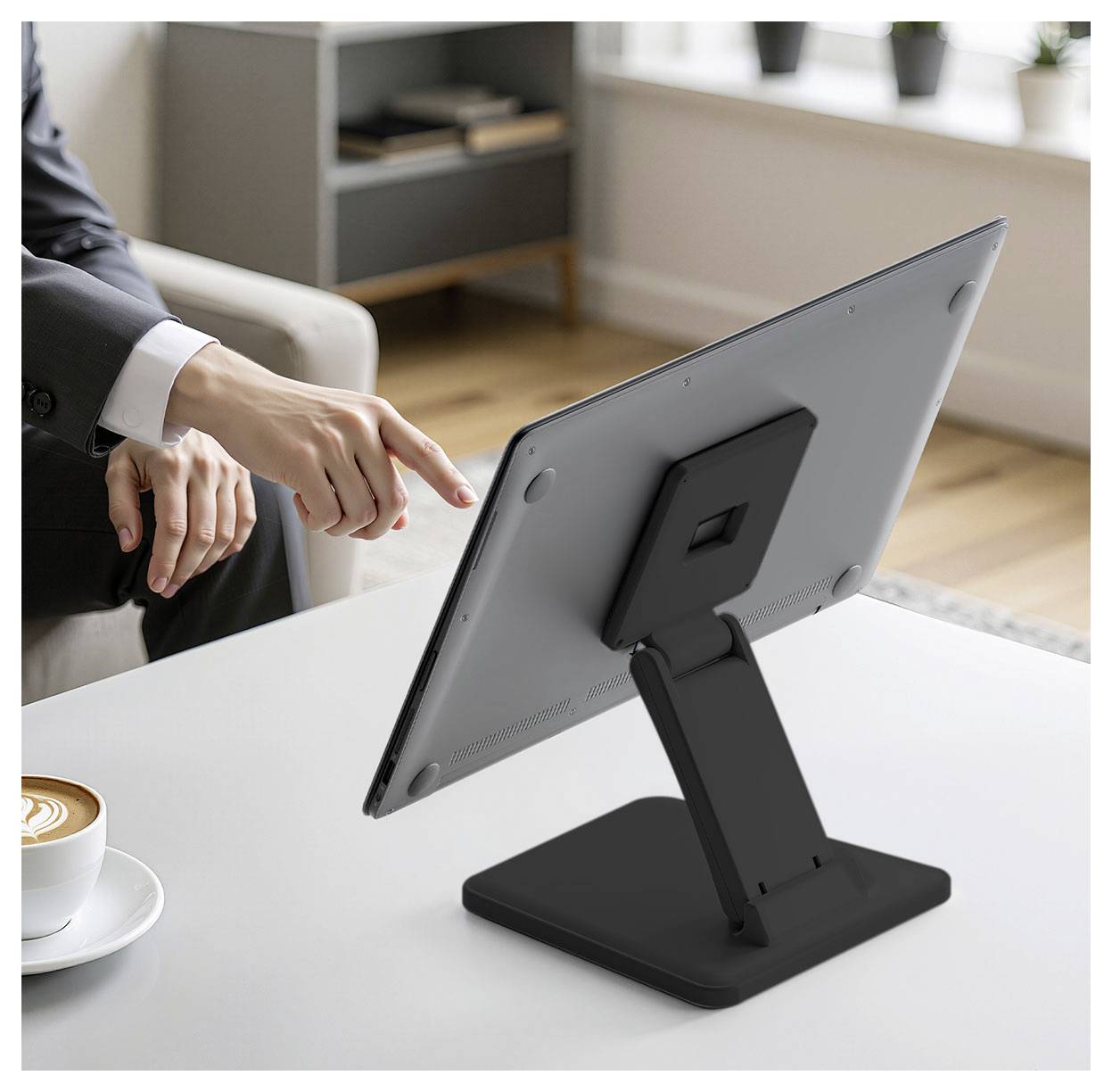 Eine Person im Anzug zeigt auf die Rückseite eines Tablets auf einem Ständer neben einer Kaffeetasse und deutet damit auf die Einrichtung oder Demonstration des Geräts hin.