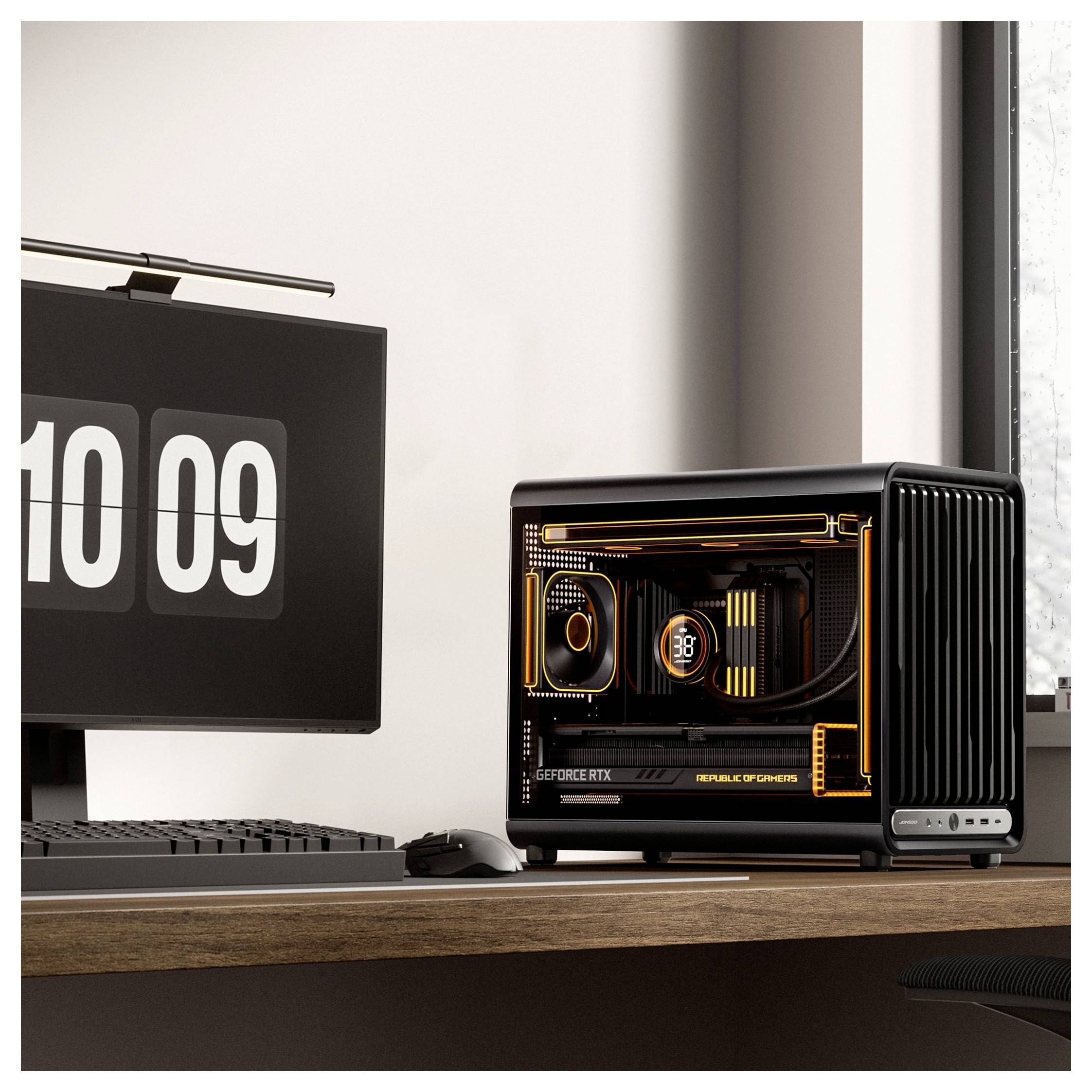 Kompakter Gaming-Desktop-PC mit durchsichtigem Seitenpaneel, orange beleuchteten Komponenten, neben einem Monitor, der '10 09' auf einem Schreibtisch anzeigt.
