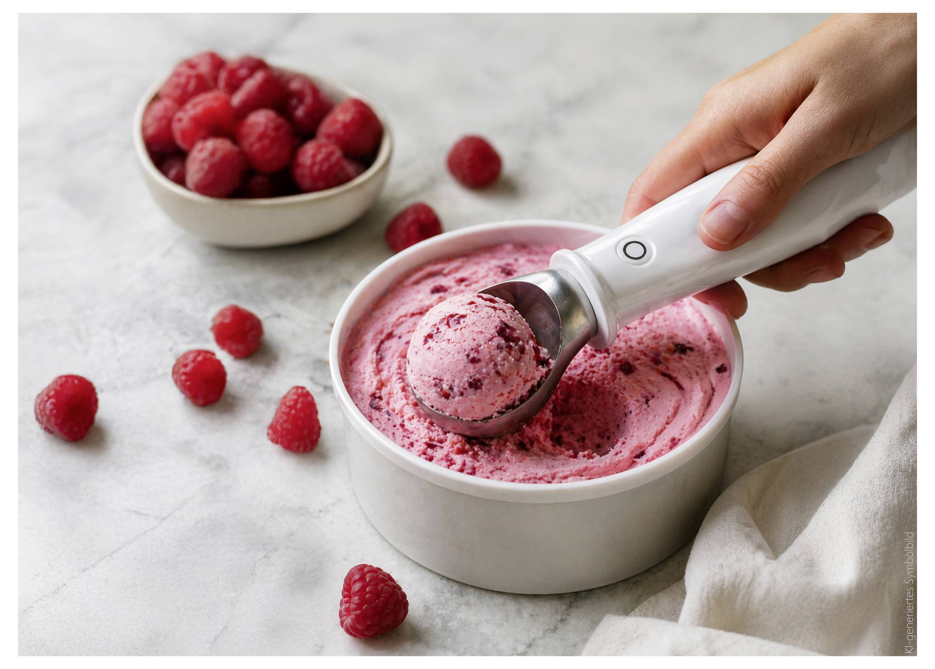 Eine Hand schöpft Himbeereis mit einem Löffel aus einer Schüssel, während frische Himbeeren in der Nähe auf einer hellen Arbeitsplatte liegen.