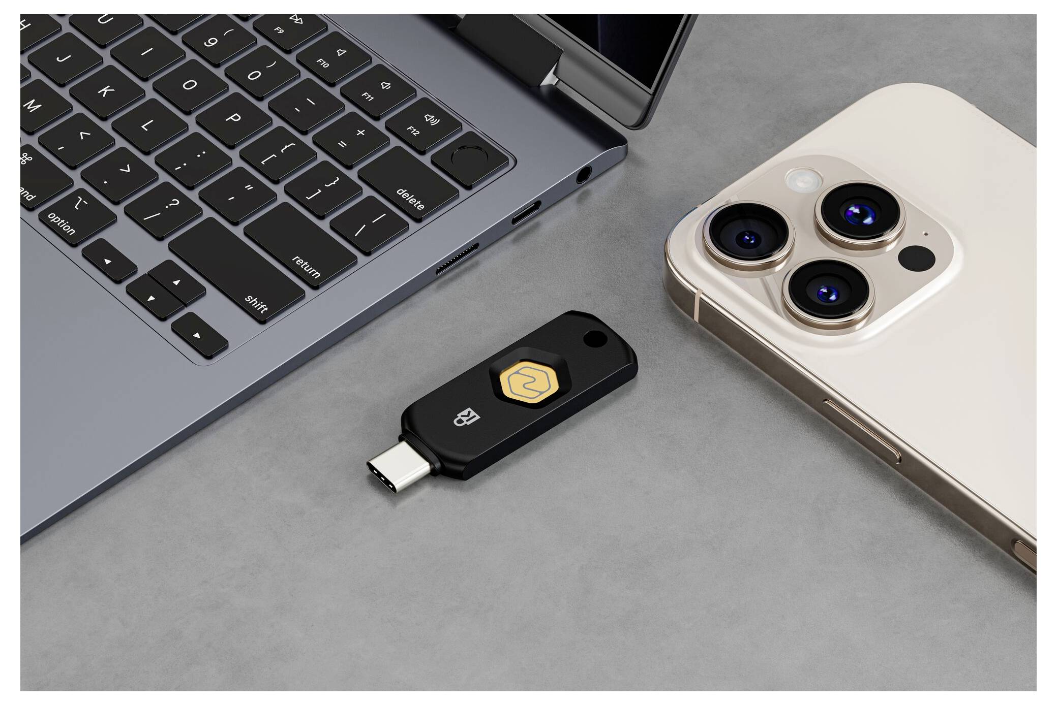 Schwarzer USB-C-Sicherheitsschlüssel zwischen einem Laptop und einem iPhone auf einer grauen Oberfläche, der Zwei-Faktor-Authentifizierungs-Hardware für die Gerätenanmeldung zeigt.