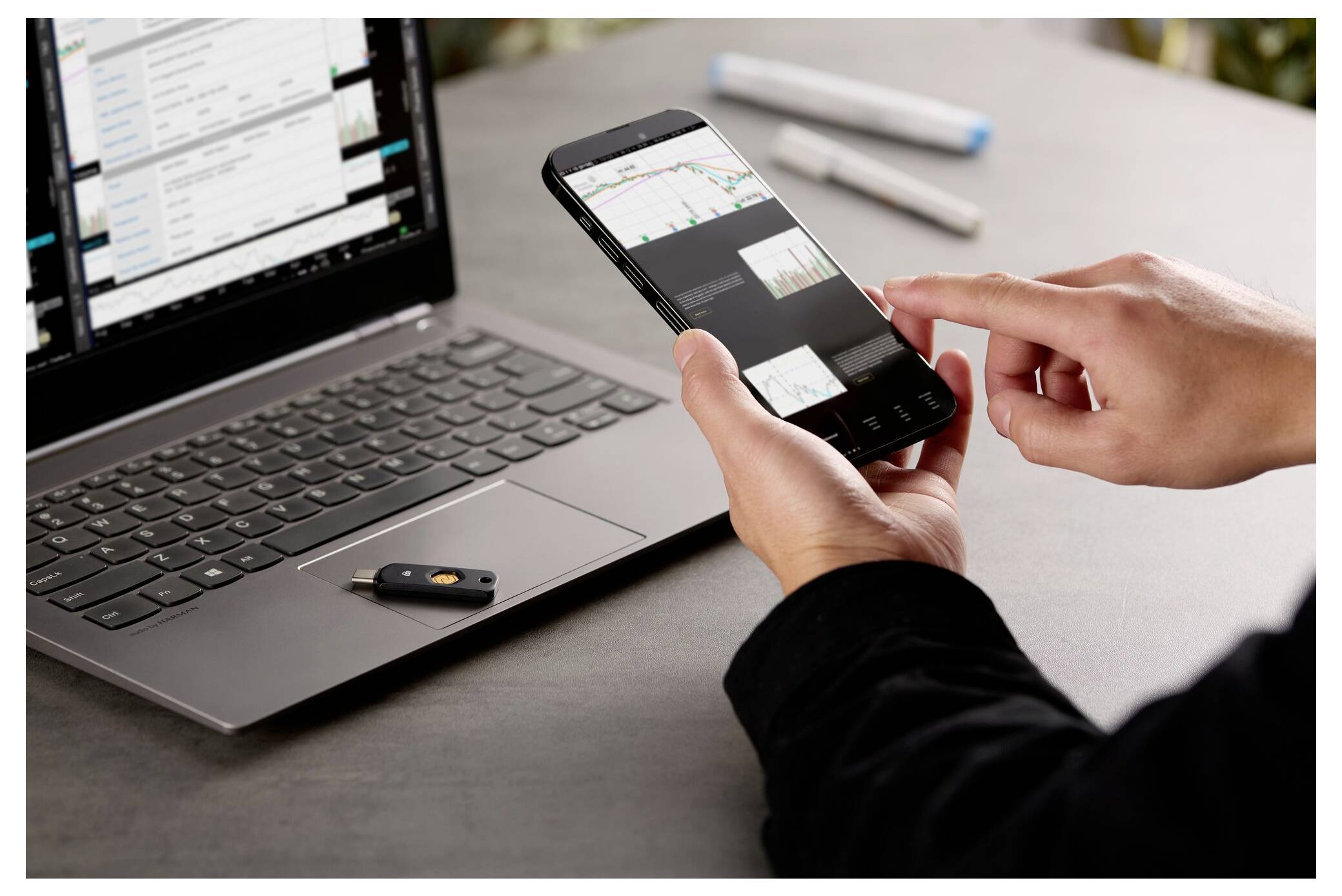 Person, die ein Smartphone mit einem Börsendiagramm überprüft, während sie an einem Laptop arbeitet, was auf mobilen Handel oder finanzielle Überwachung hindeutet.