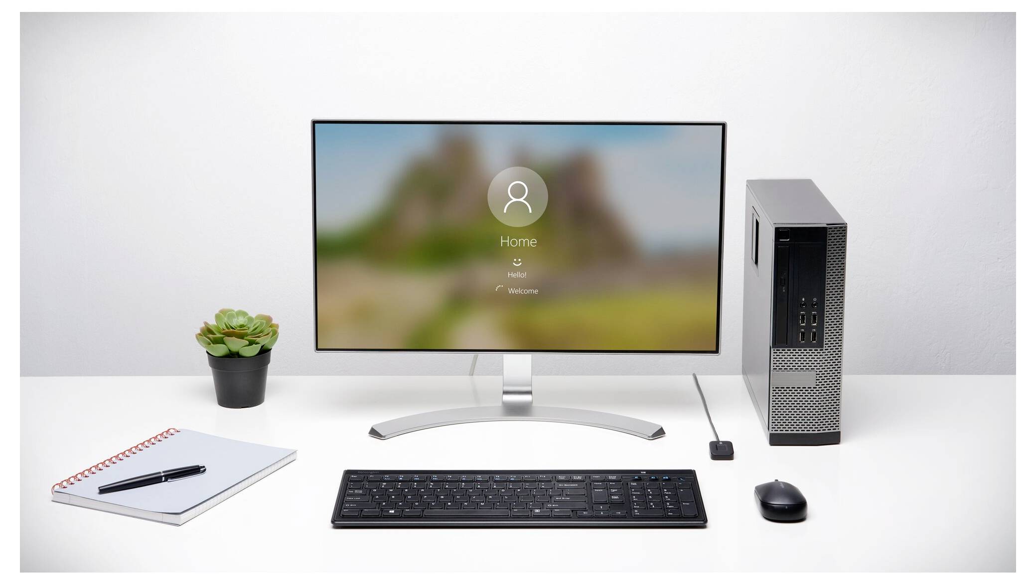 Desktop-Computer-Setup mit Monitor, der einen Anmeldebildschirm anzeigt, Tastatur, Maus, kleine Pflanze, Notizbuch und Tower-PC auf einem weißen Schreibtisch.