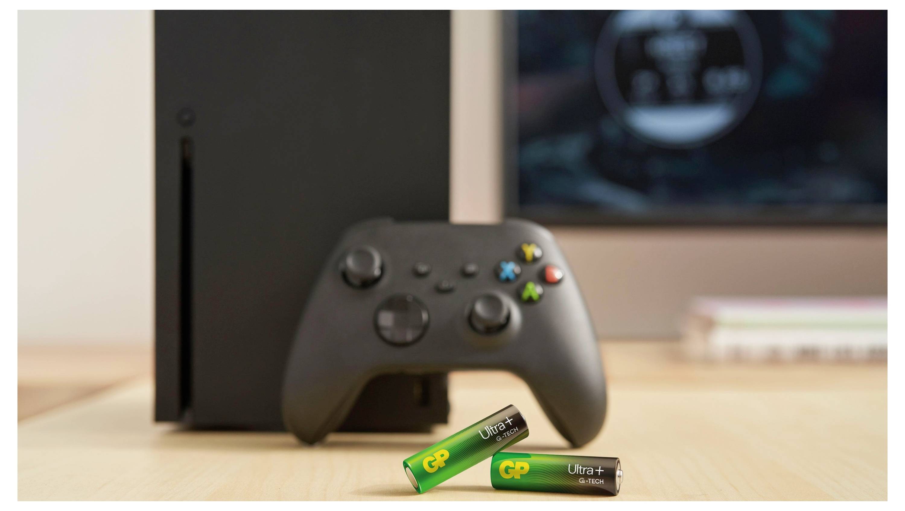 Schwarze Xbox-Konsole und Controller auf einem Schreibtisch mit zwei grünen GP Ultra-Batterien davor, was auf batteriebetriebenes Gaming-Zubehör hindeutet.