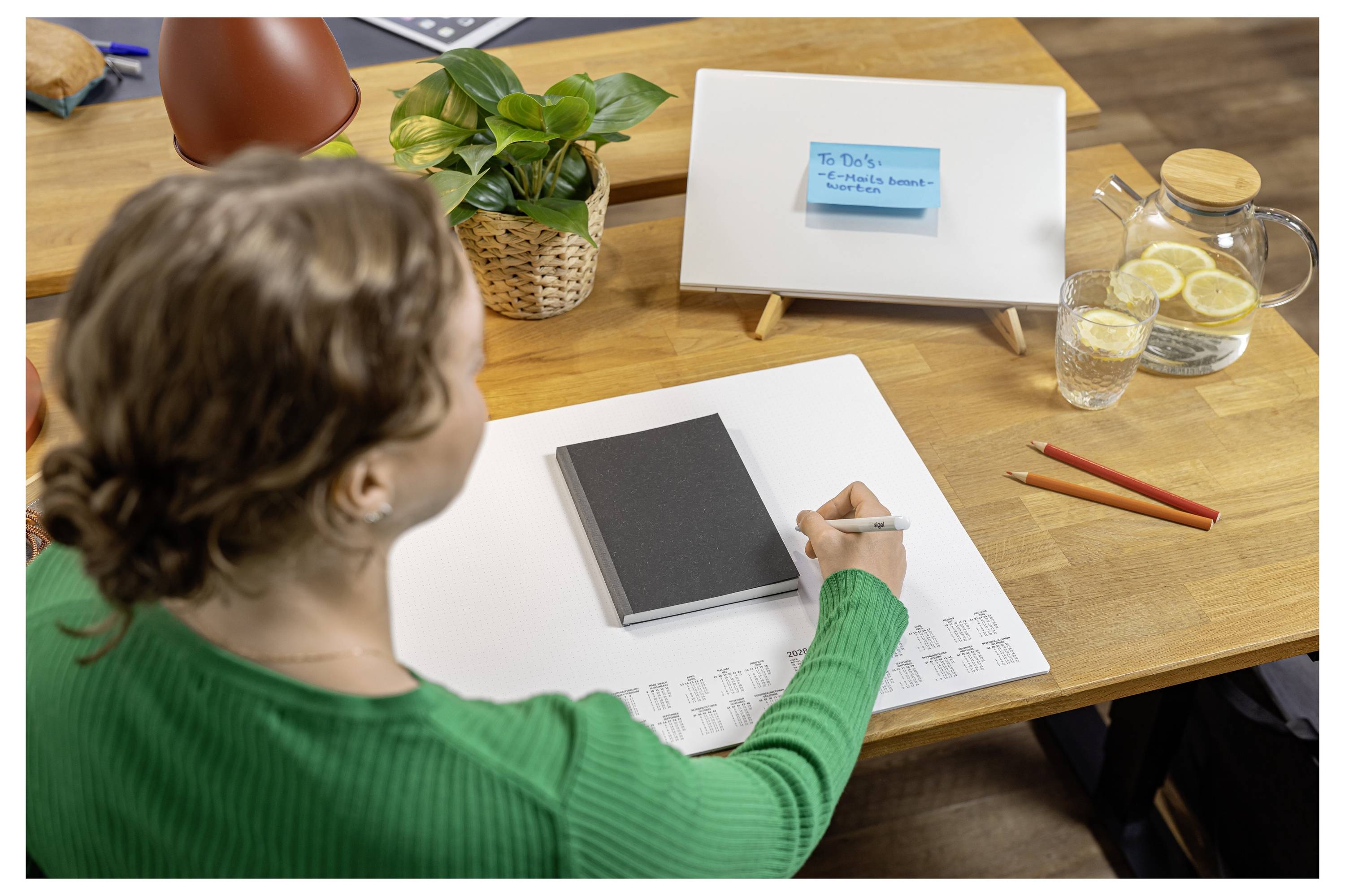 Eine Person in einem grünen Pullover schreibt in einem Notizbuch an einem Schreibtisch mit einer kleinen Whiteboard-Notiz, Stiften, einer Pflanze und einer Kanne mit Zitronenwasser.