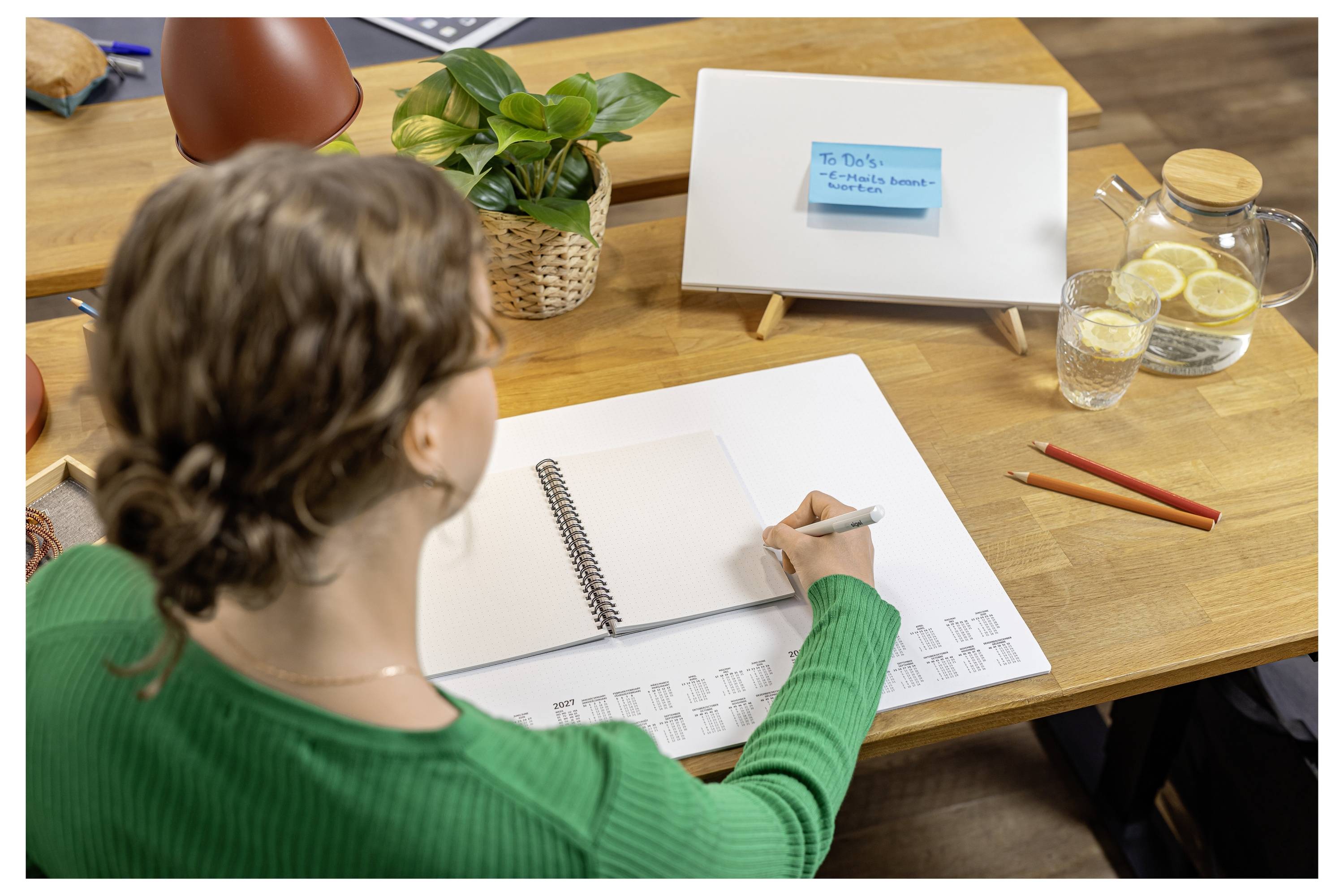 Person an einem Schreibtisch, der in ein Notizbuch schreibt, während er auf ein kleines Whiteboard mit einer blauen Haftnotiz schaut, was auf Planung oder Brainstorming hindeutet.