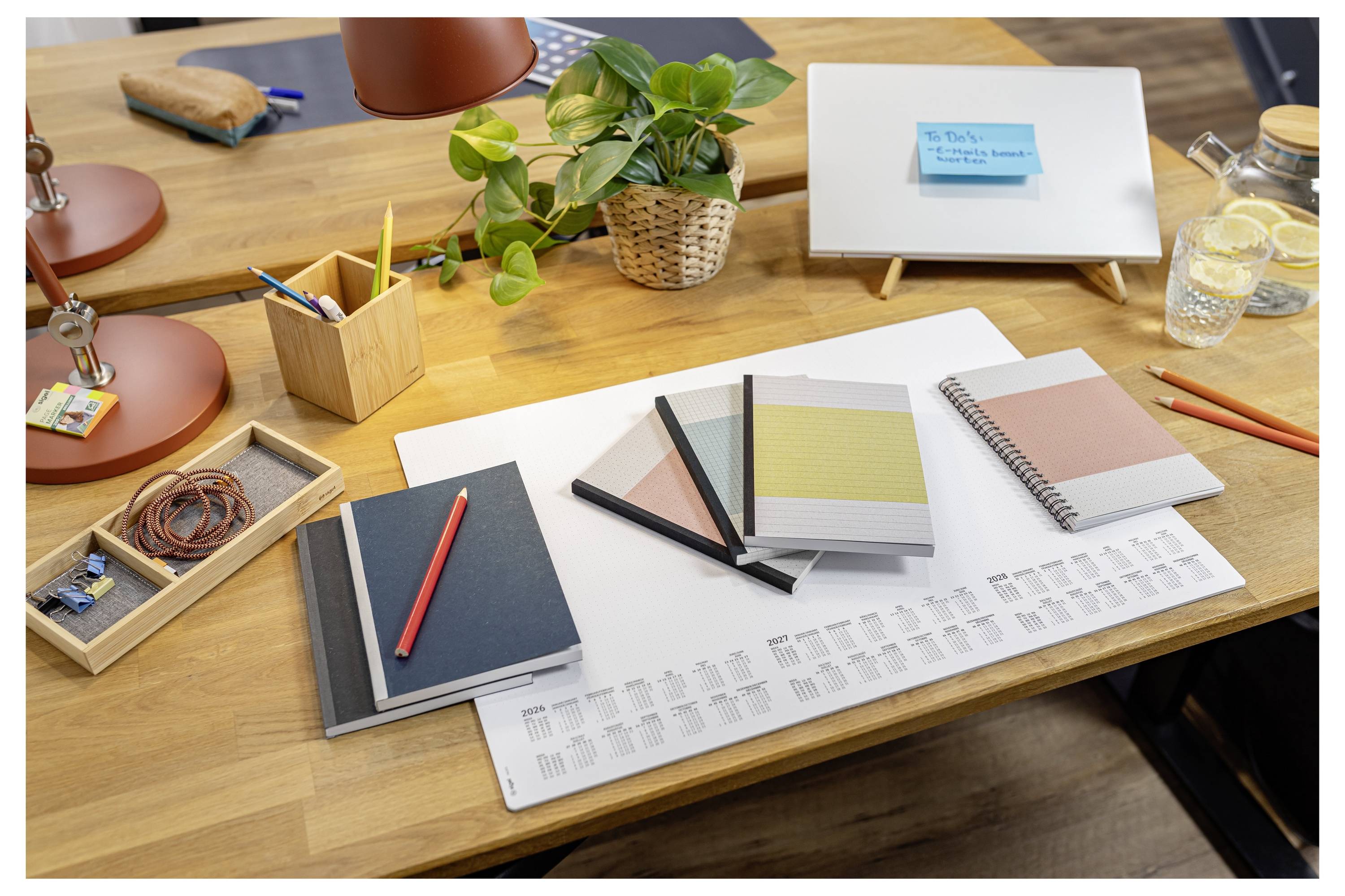 Arbeitsplatz-Schreibtisch mit Notizbüchern, Stiften, Kalender, Topfpflanze, Lampen und einem kleinen Whiteboard mit einer blauen Haftnotiz.