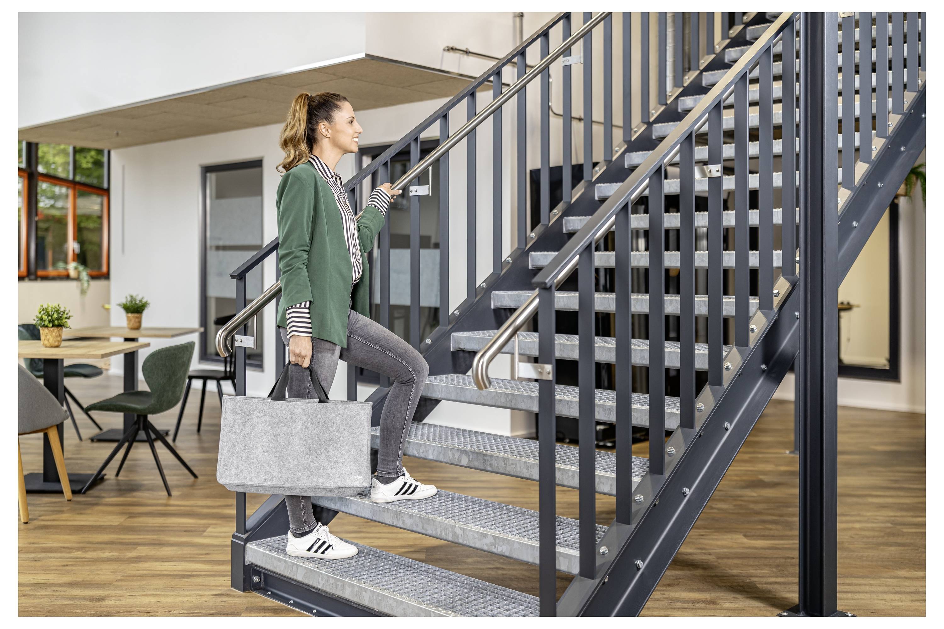 Eine Frau, die eine graue Tragetasche trägt, erklimmt eine Innentreppe aus Metall in einem modernen Büro- oder Wohnungsinterieur.