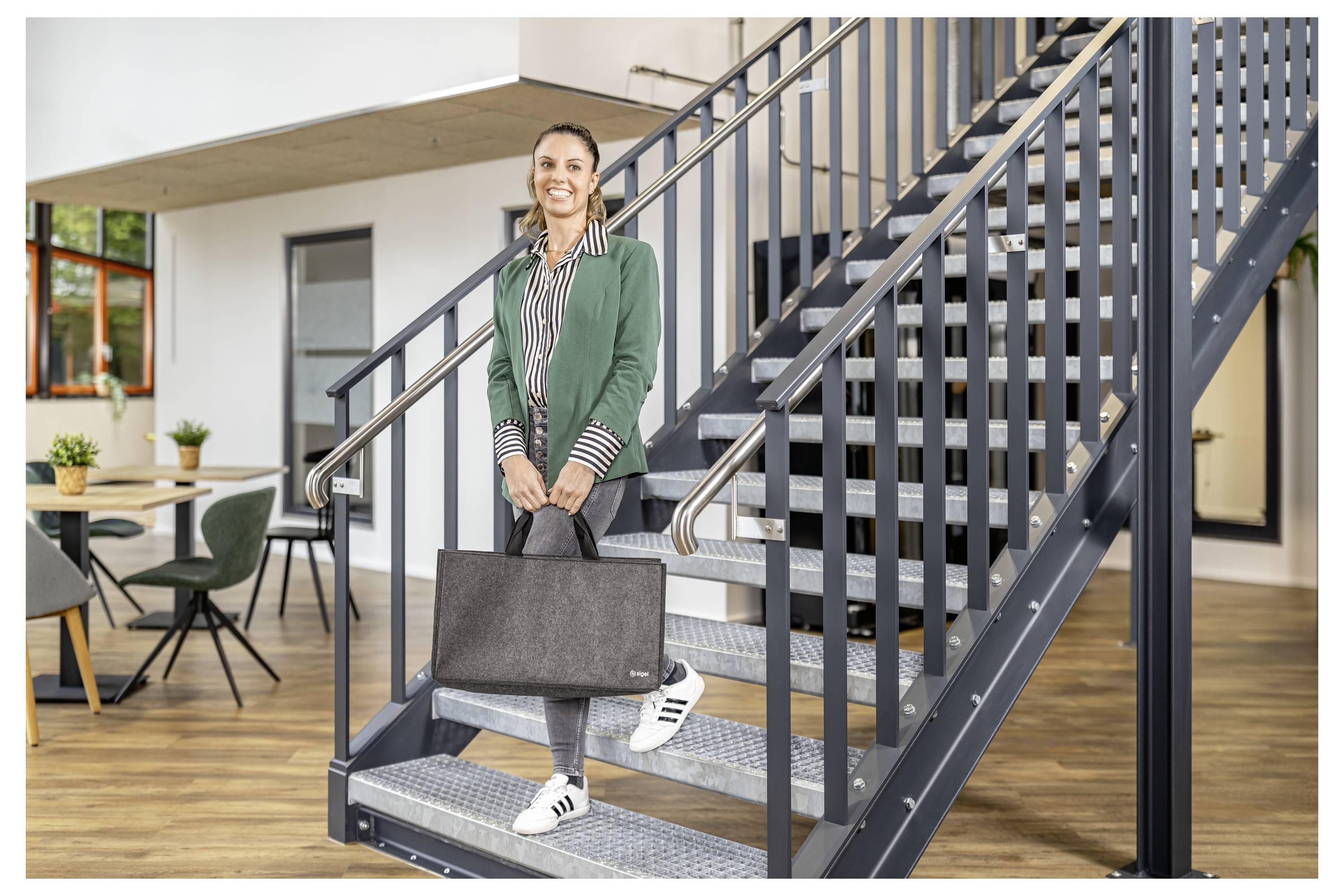 Lächelnde Frau, die einen Aktenkoffer hält, steht auf einer Innentreppe in einem modernen Bürogebäude.