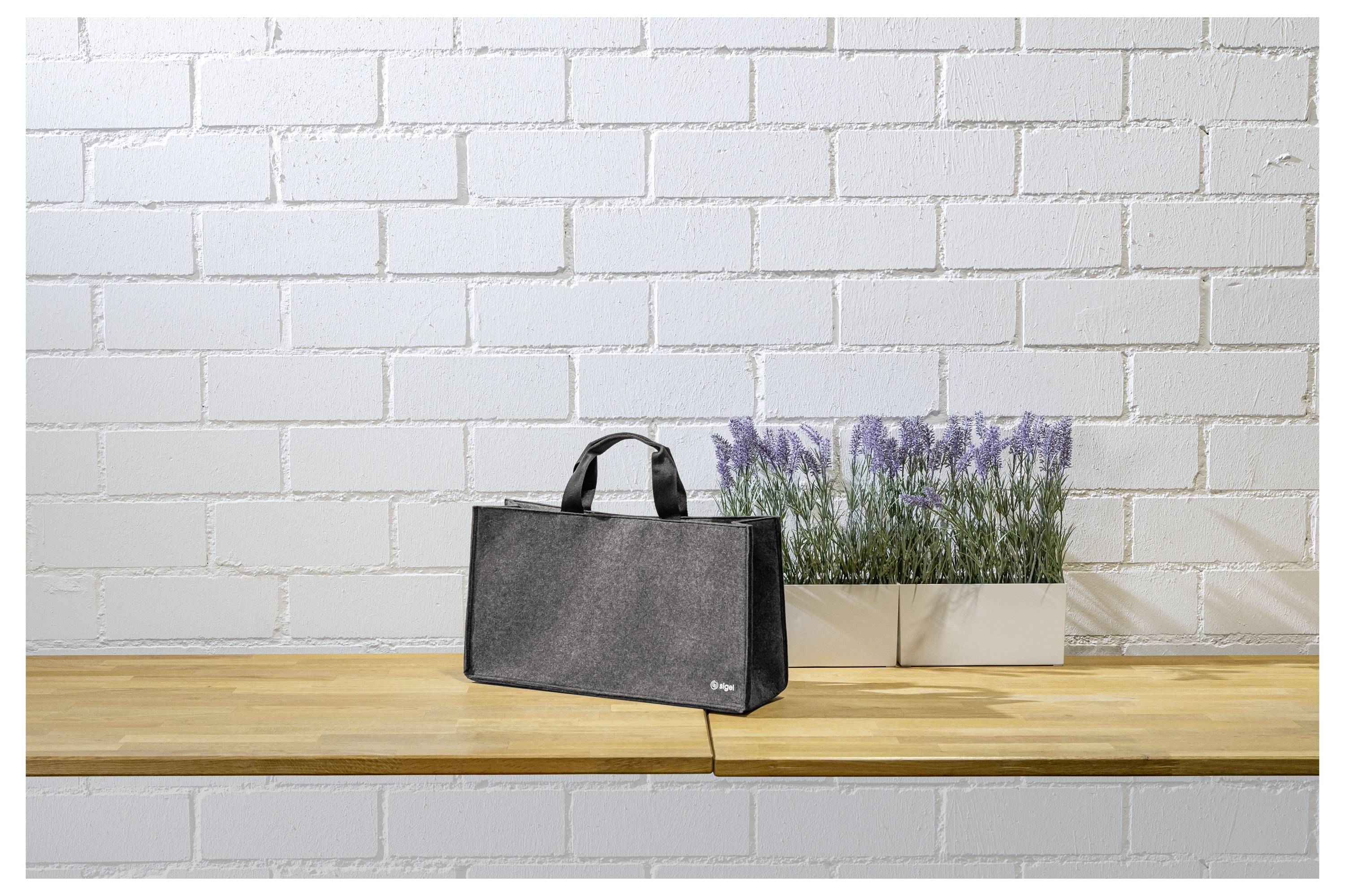 Schwarze Tragetasche auf einem Holztisch neben einem weißen Blumenkübel mit violetten Blüten vor einer weißen Ziegelmauer.