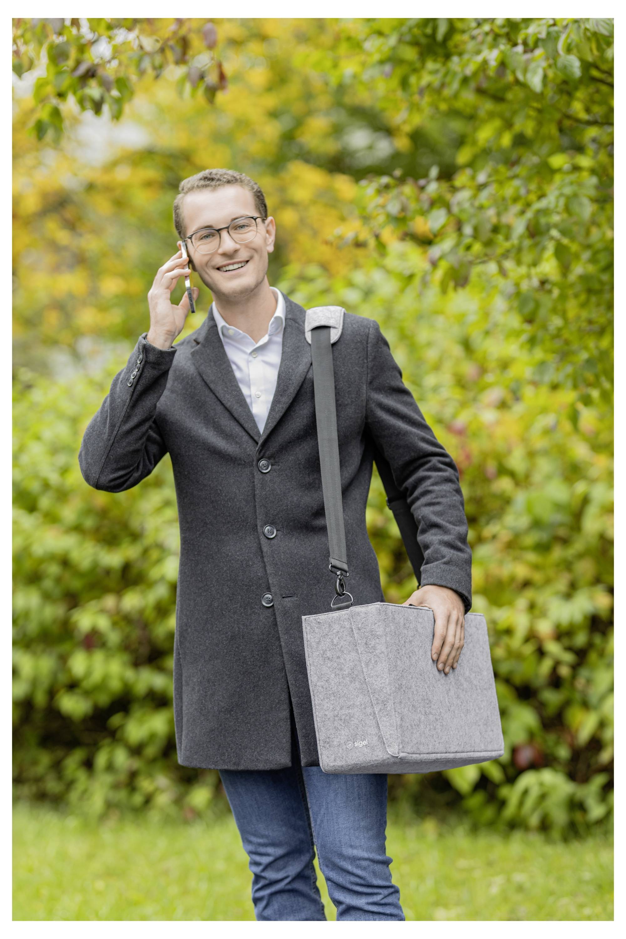 Ein lächelnder Mann mit Brille steht draußen in einem Park und telefoniert, während er eine graue Laptop-Tasche hält.