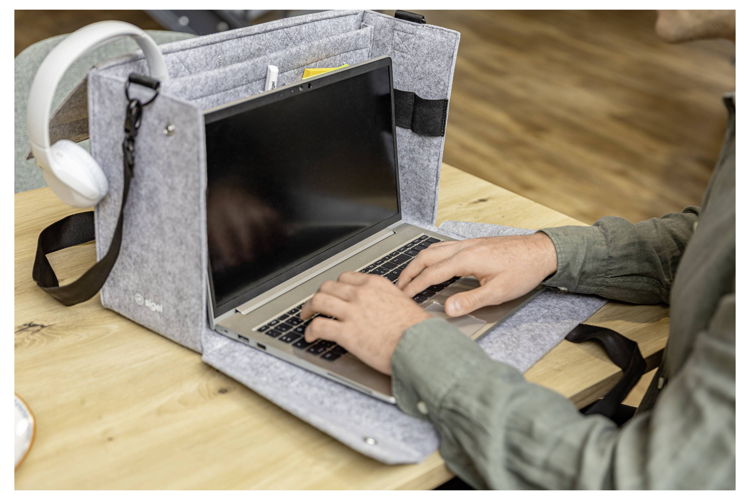 Person tippt auf einem Laptop in einem grauen Filz-Tragetasche auf einem Holztisch, mit weißen Kopfhörern, die daneben hängen.