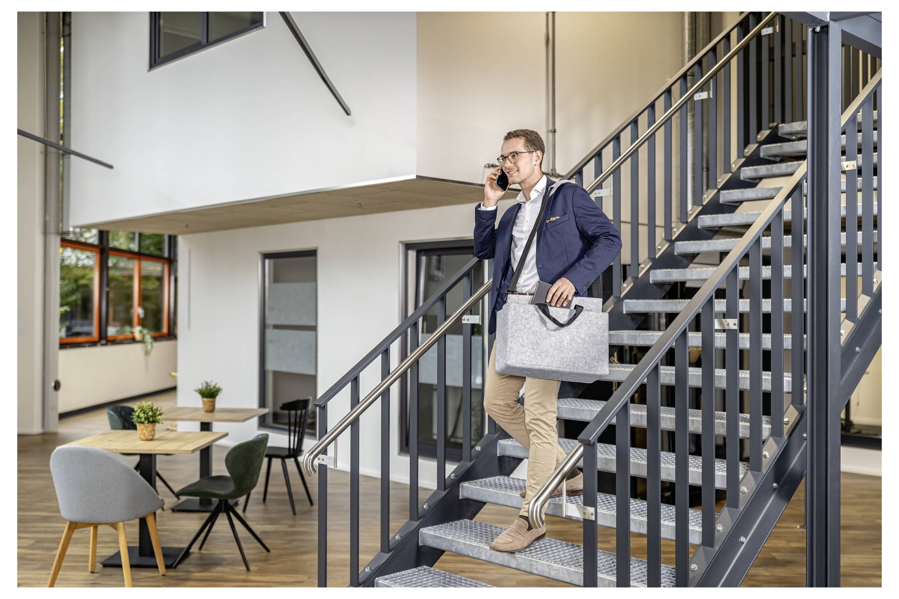 Geschäftsmann in einem Blazer steigt eine Bürotreppe hinab, während er am Telefon spricht und eine Laptoptasche in einem modernen Interieur trägt.