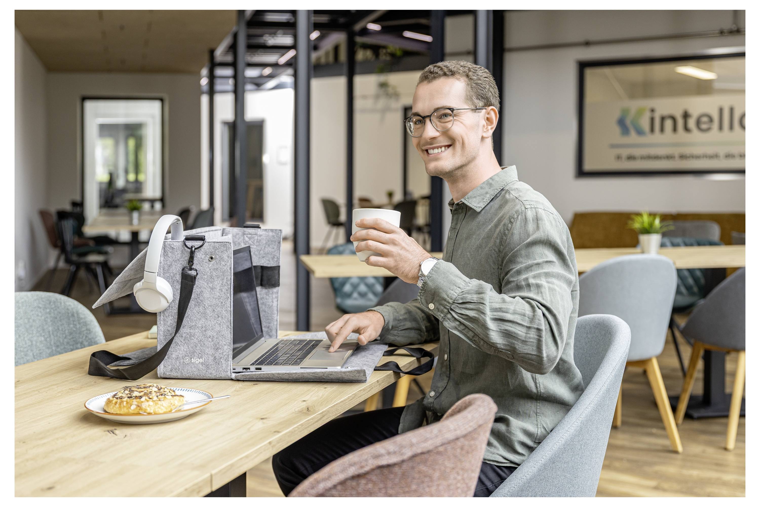 Ein Mann in einem Coworking Space trinkt Kaffee, während er an einem Laptop an einem gemeinsamen Tisch arbeitet, mit einem Gebäck und Bürotaschen in der Nähe.