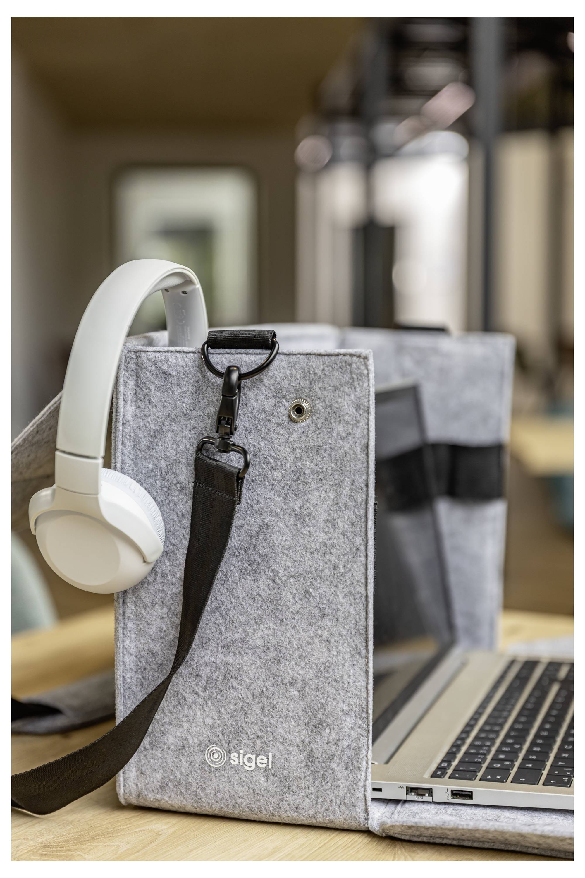 Eine graue Filz-Laptophülle mit einem weißen Headset und schwarzem Riemen, neben einem aufgeklappten Laptop auf einem Holzschreibtisch.