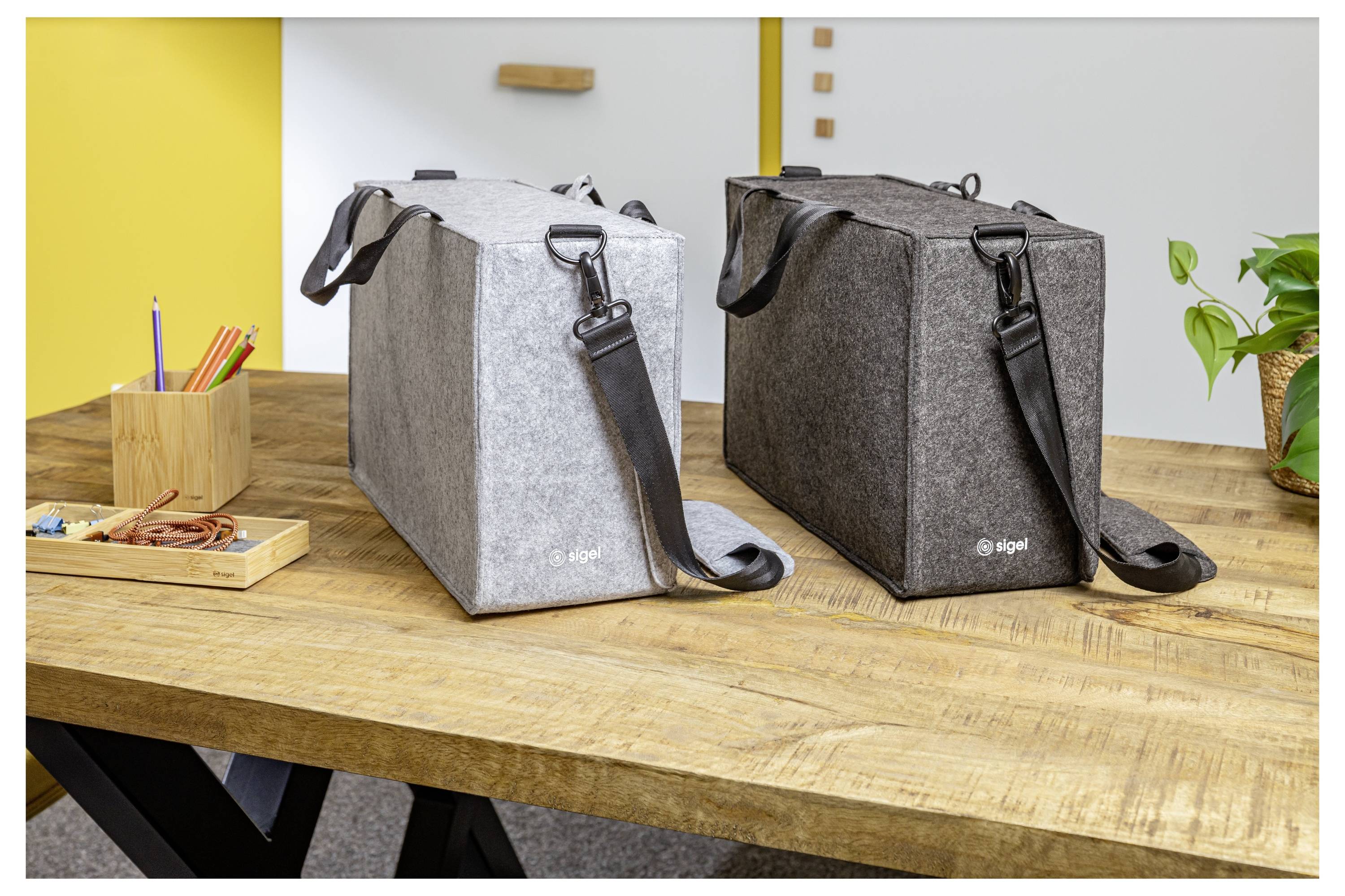 Zwei isolierte Schultertaschen, eine hellgrau und eine dunkelgrau, stehen auf einem Holztisch in einer büroähnlichen Umgebung.