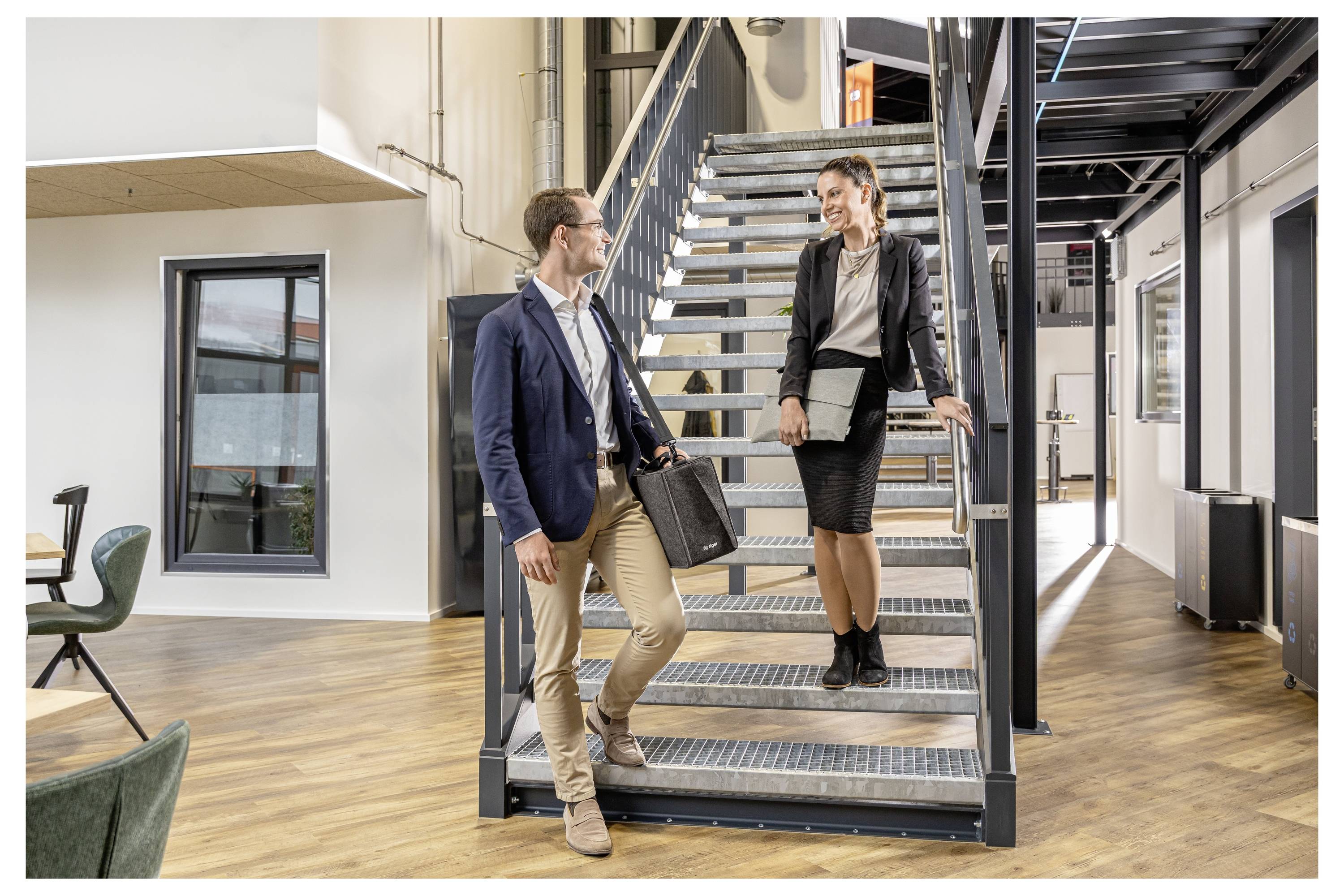 Zwei Geschäftskollegen unterhalten sich auf einer Bürotreppe in einem modernen Arbeitsraum, mit Schreibtischen und großen Fenstern in der Nähe.
