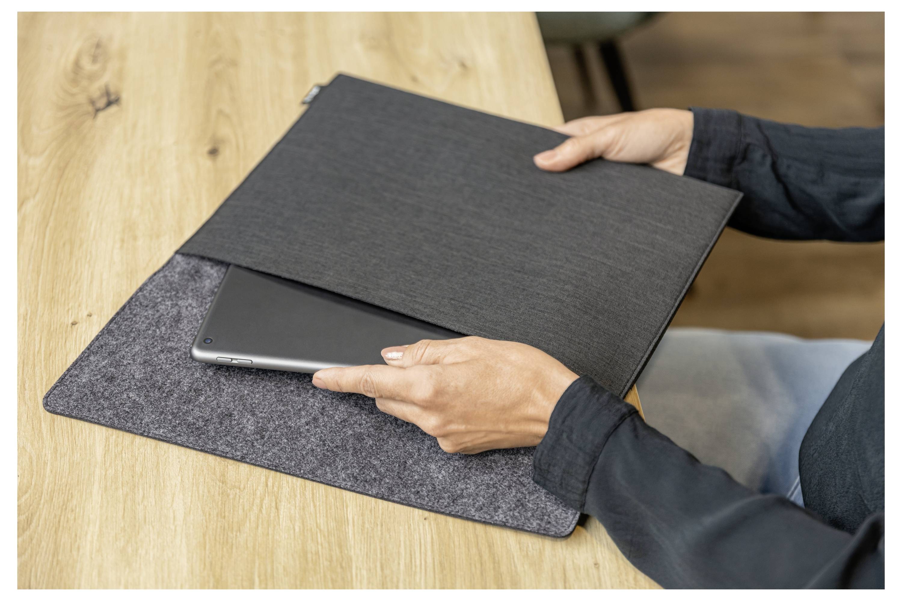 Eine Person schiebt einen Laptop in eine dunkle Filzhülle auf einem Holztisch.