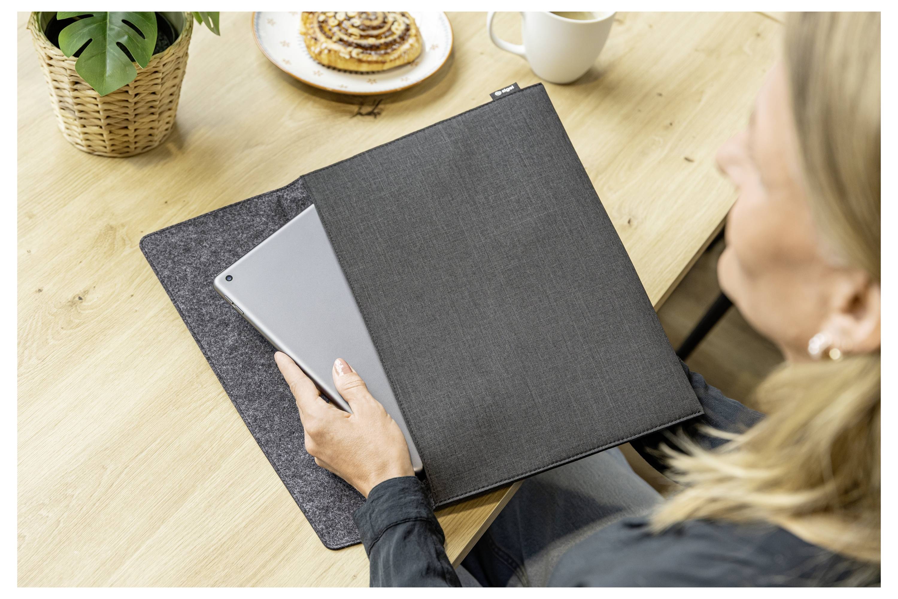 Frau an einem Tisch benutzt ein Tablet neben einem geschlossenen schwarzen Ordner, mit Kaffee und einem Gebäck auf dem Schreibtisch.