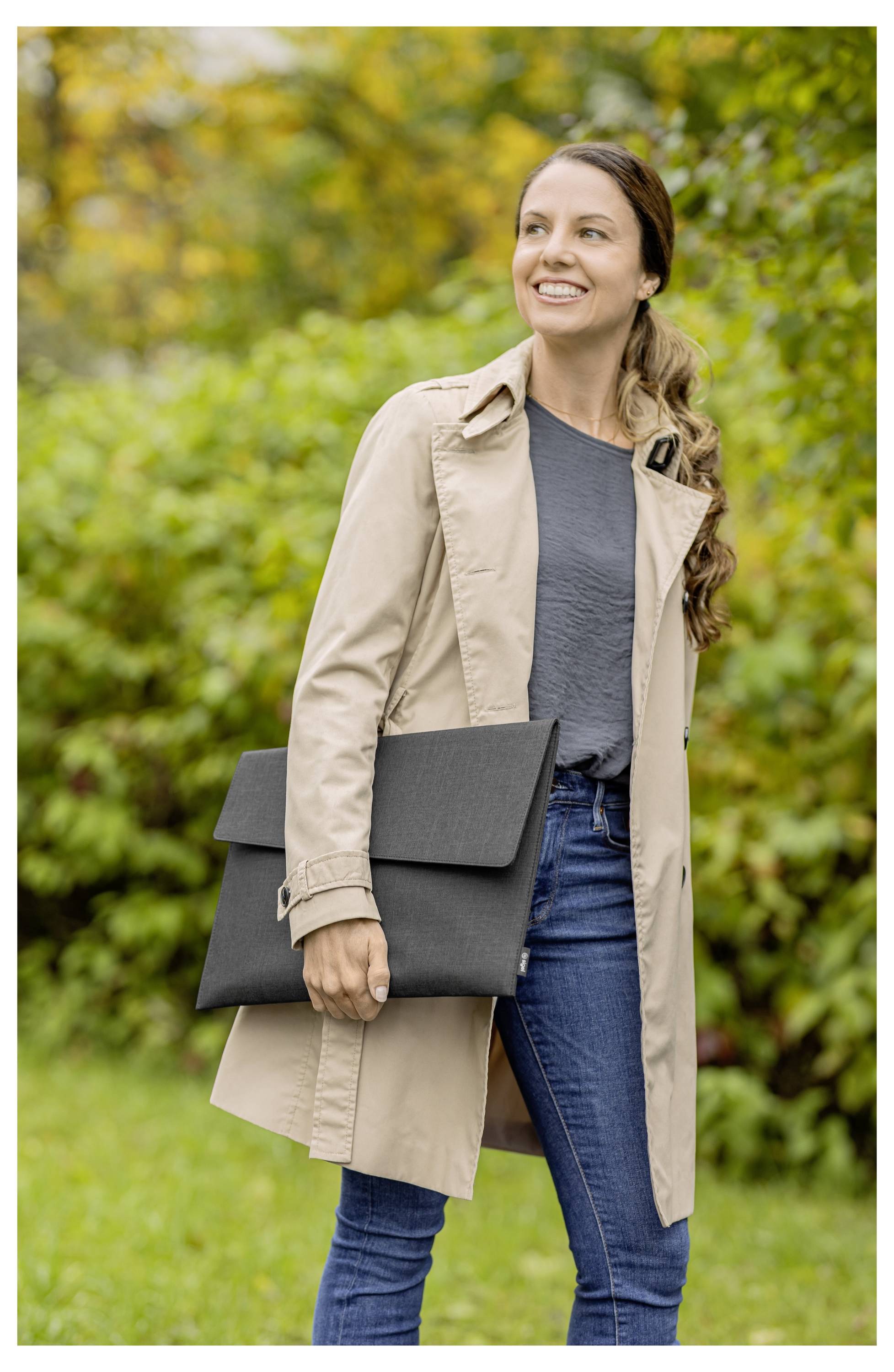 Lächelnde Frau im Freien in einem beigen Mantel und Jeans, die eine schwarze Laptop-Tasche vor einem unscharfen grünen Herbsthintergrund hält.