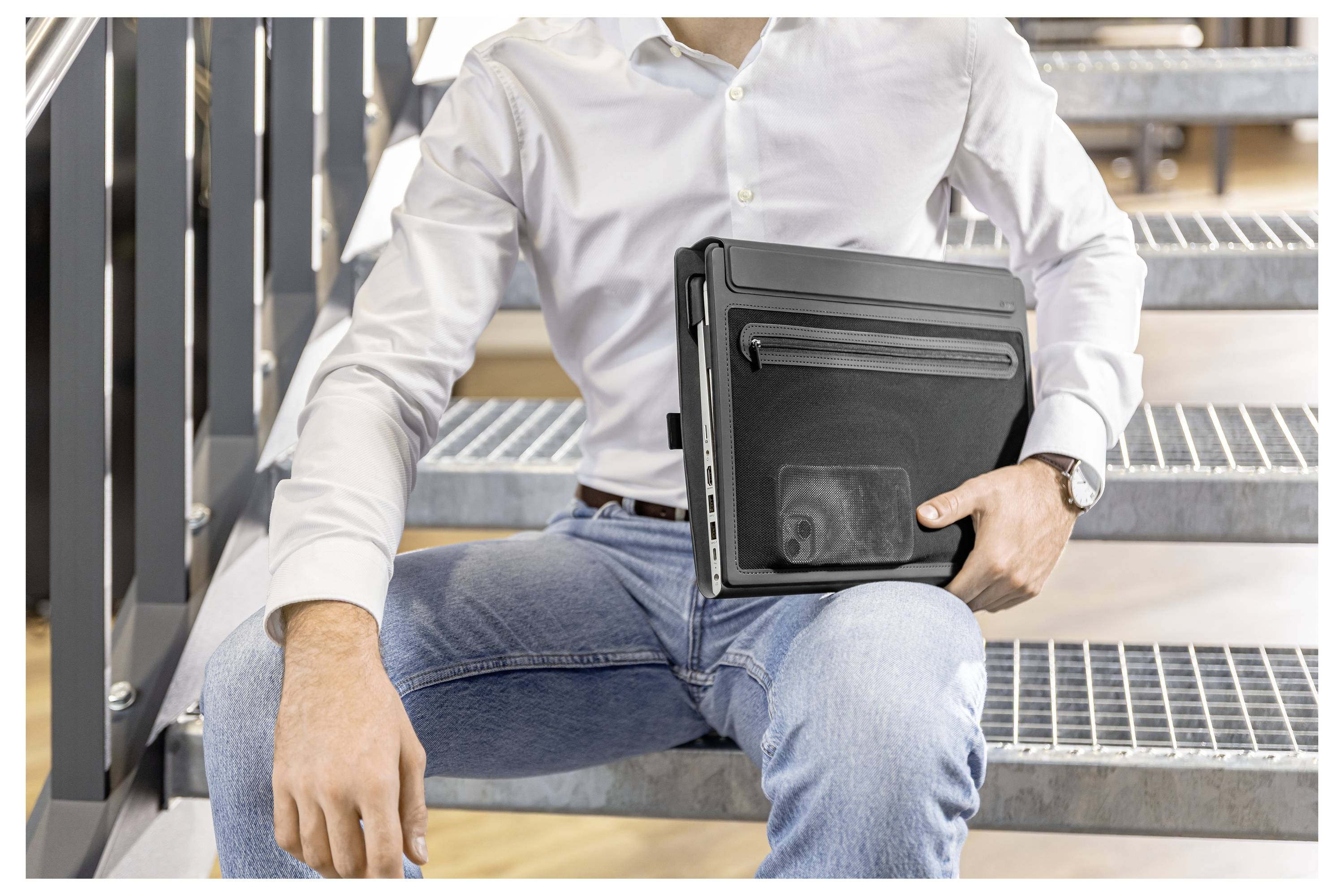 Mann, der einen geschlossenen Laptop auf Außenmetalltreppe hält, trägt ein weißes Hemd und Jeans.