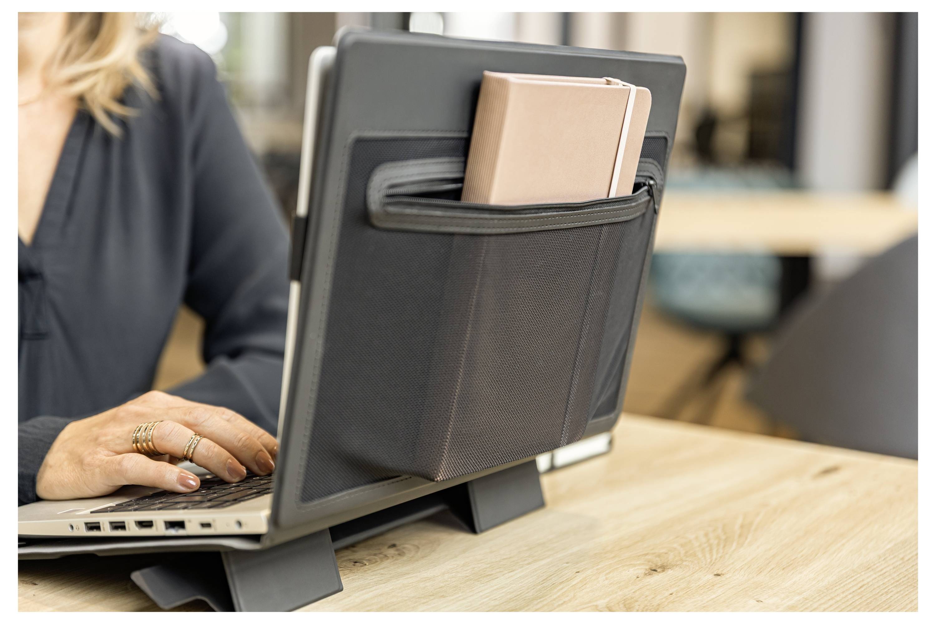 Ein Laptop auf einem Schreibtisch mit einem Mesh-Organizer, der an seiner Rückseite befestigt ist und Papiere hält, während eine Person daneben tippt.