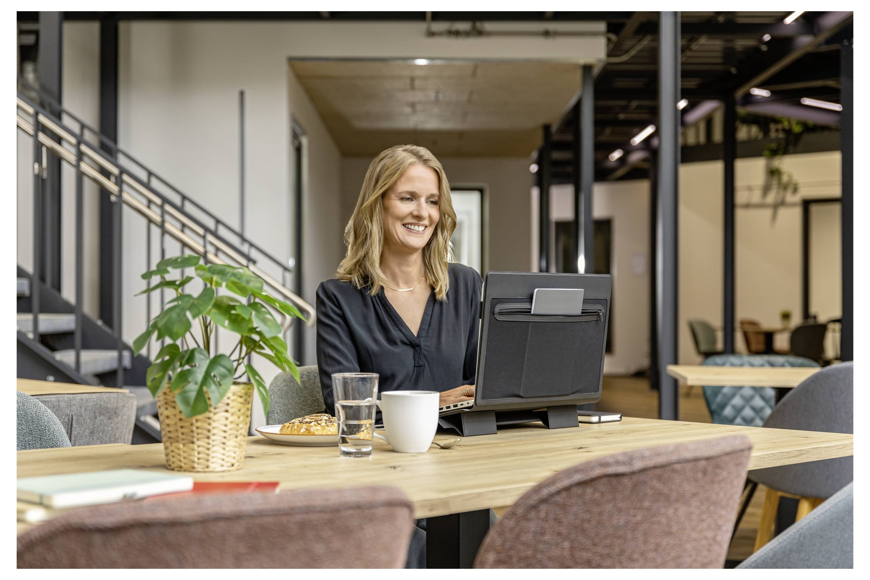 Lächelnde Frau arbeitet an einem Laptop an einem Tisch in einer modernen Bürolounge, mit Kaffee, Wasser und einer Pflanze in der Nähe.