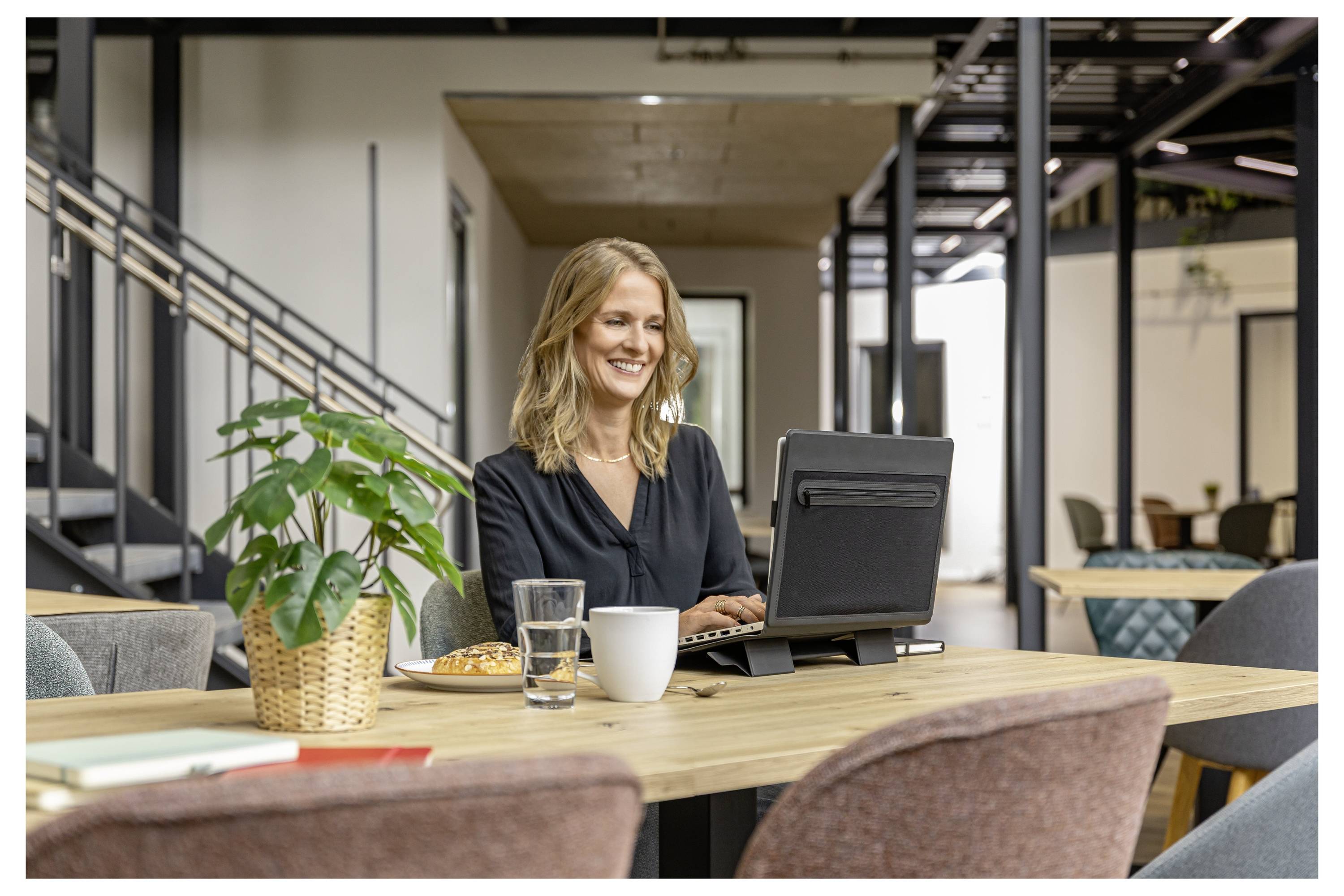 Eine Frau lächelt, während sie an einem Laptop an einem Tisch in einer modernen Bürolounge arbeitet, mit Kaffee und einem Glas Wasser in der Nähe.