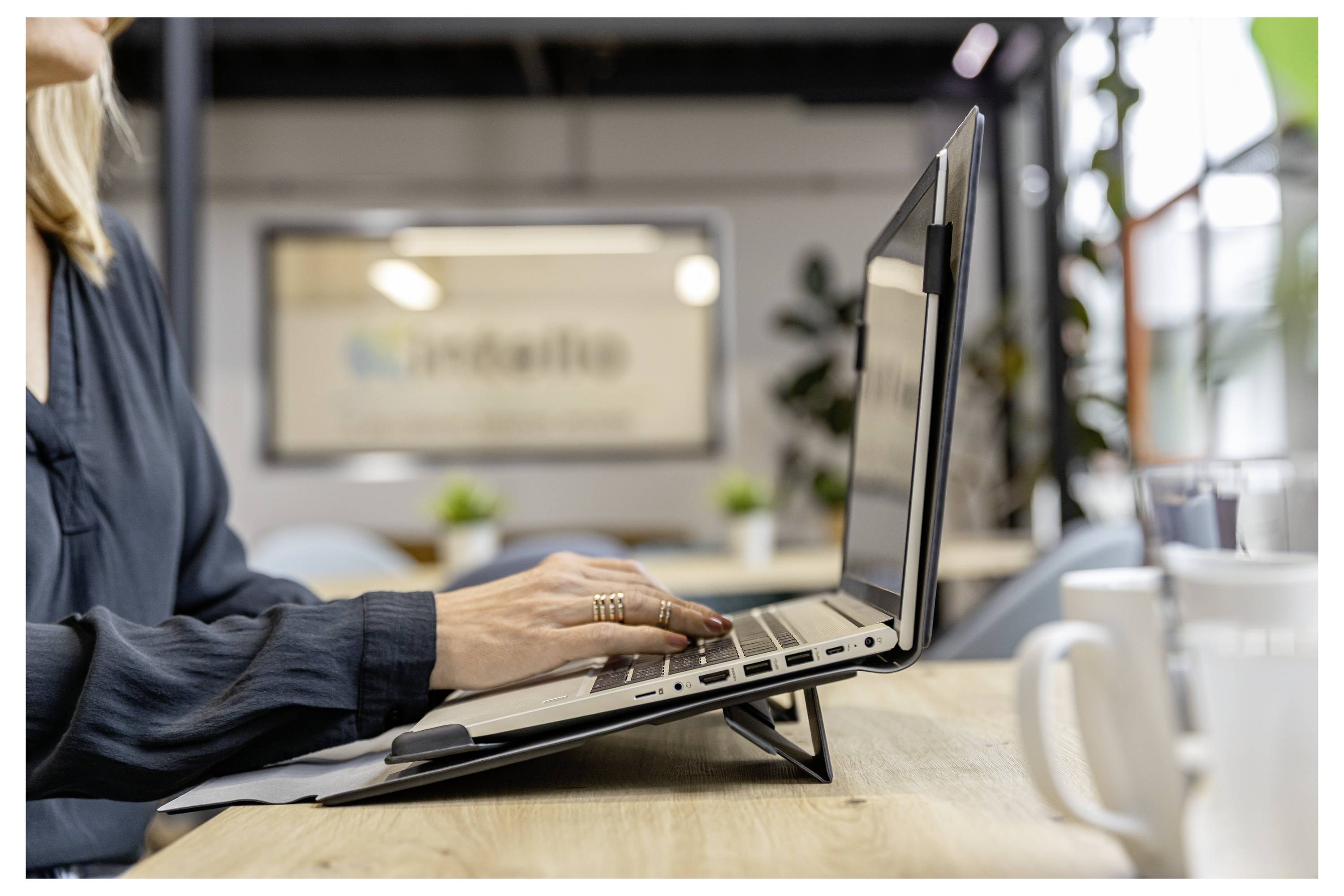 Frau, die an einem Laptop an einem Schreibtisch in einer hellen Büro- oder Café-Umgebung tippt, mit einer Tasse in der Nähe.