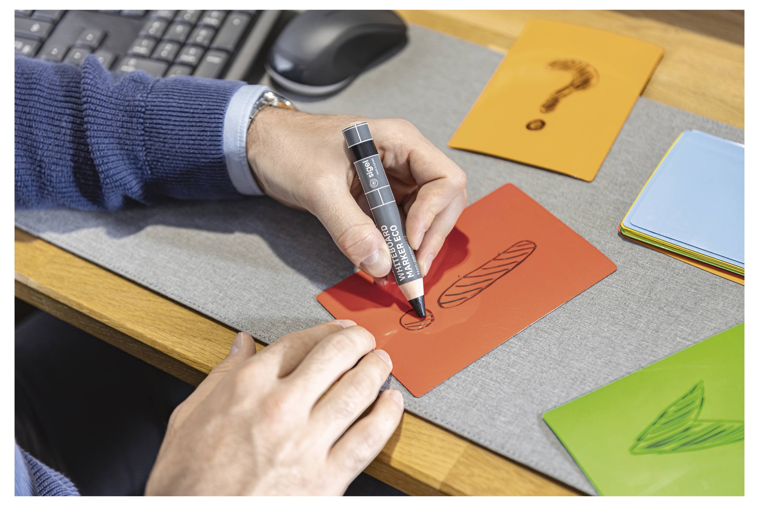 Hände zeichnen mit einem schwarzen Marker einen kleinen Kreis auf eine rote Karte an einem Schreibtisch, während in der Nähe andere farbige Karten mit Skizzenentwürfen sichtbar sind.