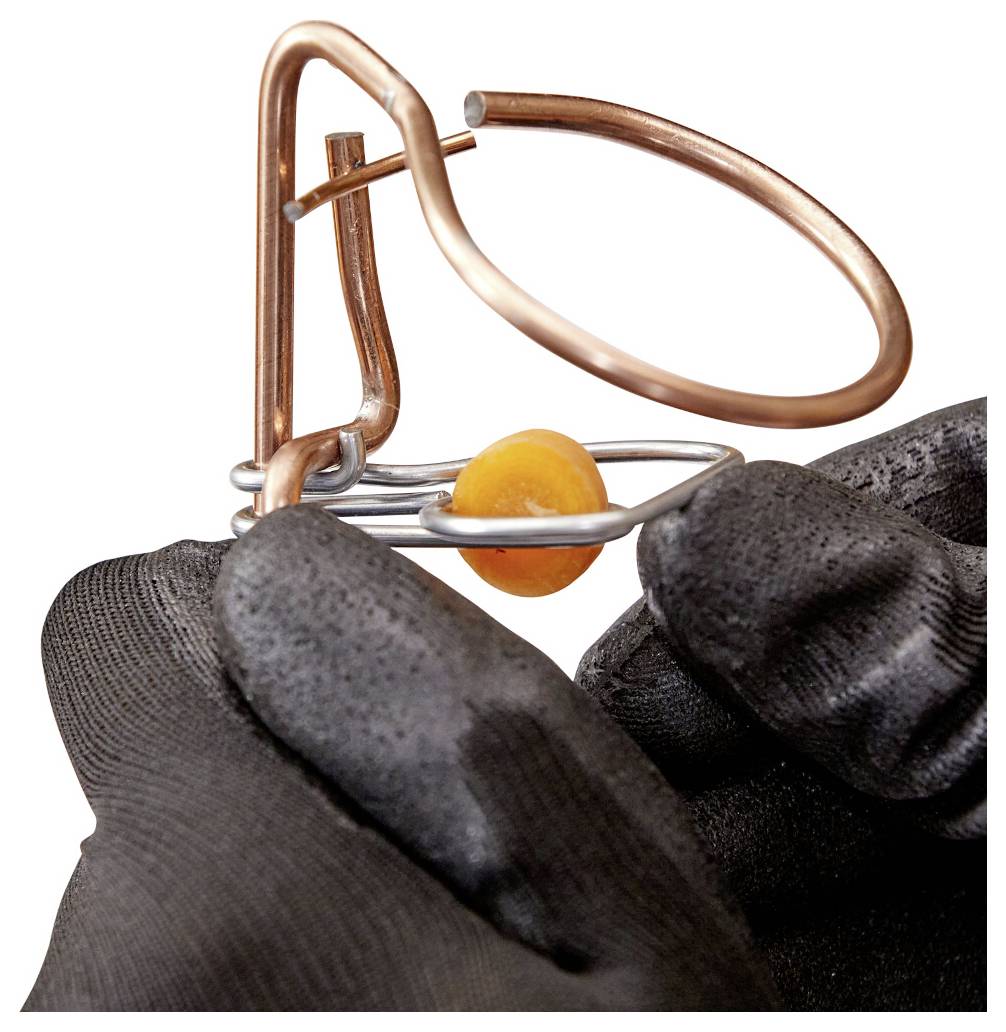 Behandschuhte Hände, die eine Kupfer- und Silberdrahtfalle mit einer orangefarbenen Perle halten, deuten auf eine Nahaufnahme von Schmuckherstellung oder Drahthandwerk hin.